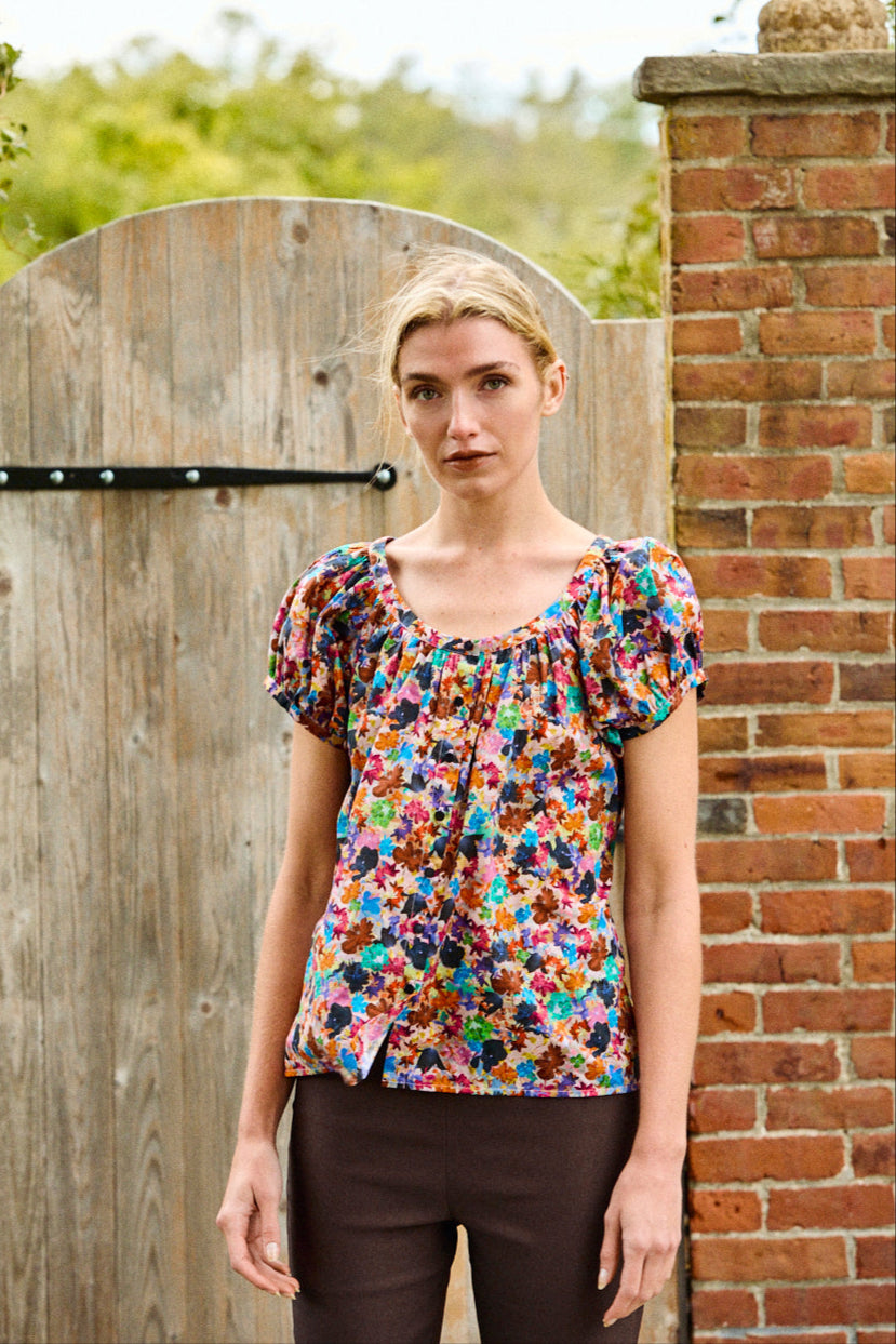 Woman wearing a colorful floral blouse standing in front of a wooden gate and brick wall.