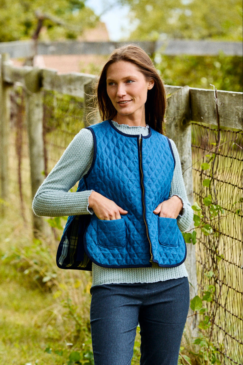 Woman wearing a blue quilted vest standing in a grassy field with a wooden fence in the background.