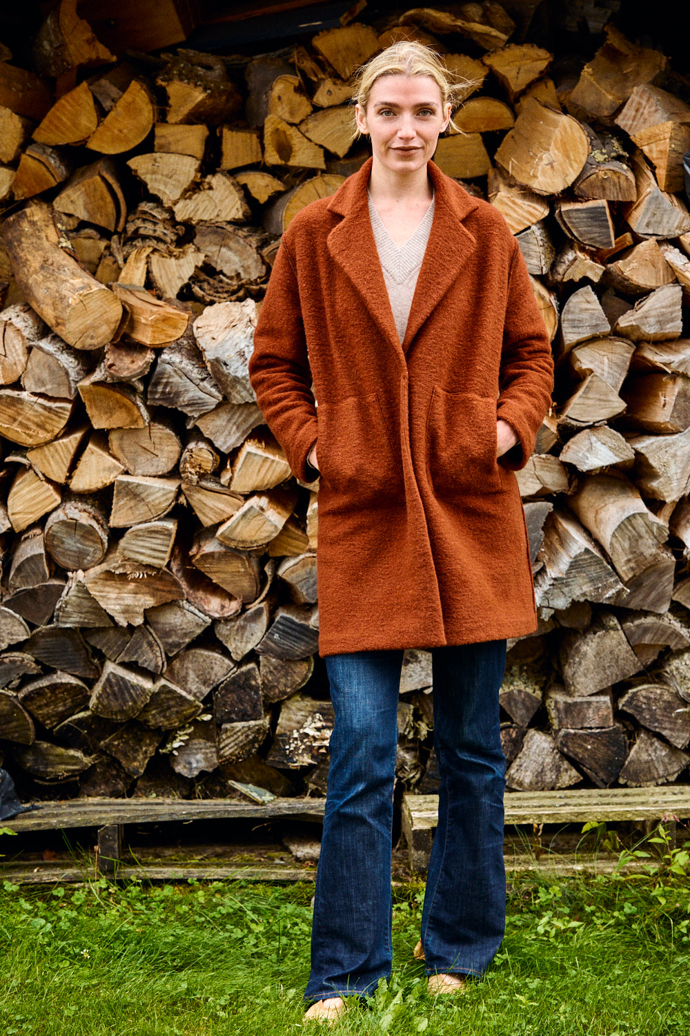 Woman wearing a brown coat standing in front of stacked logs.
