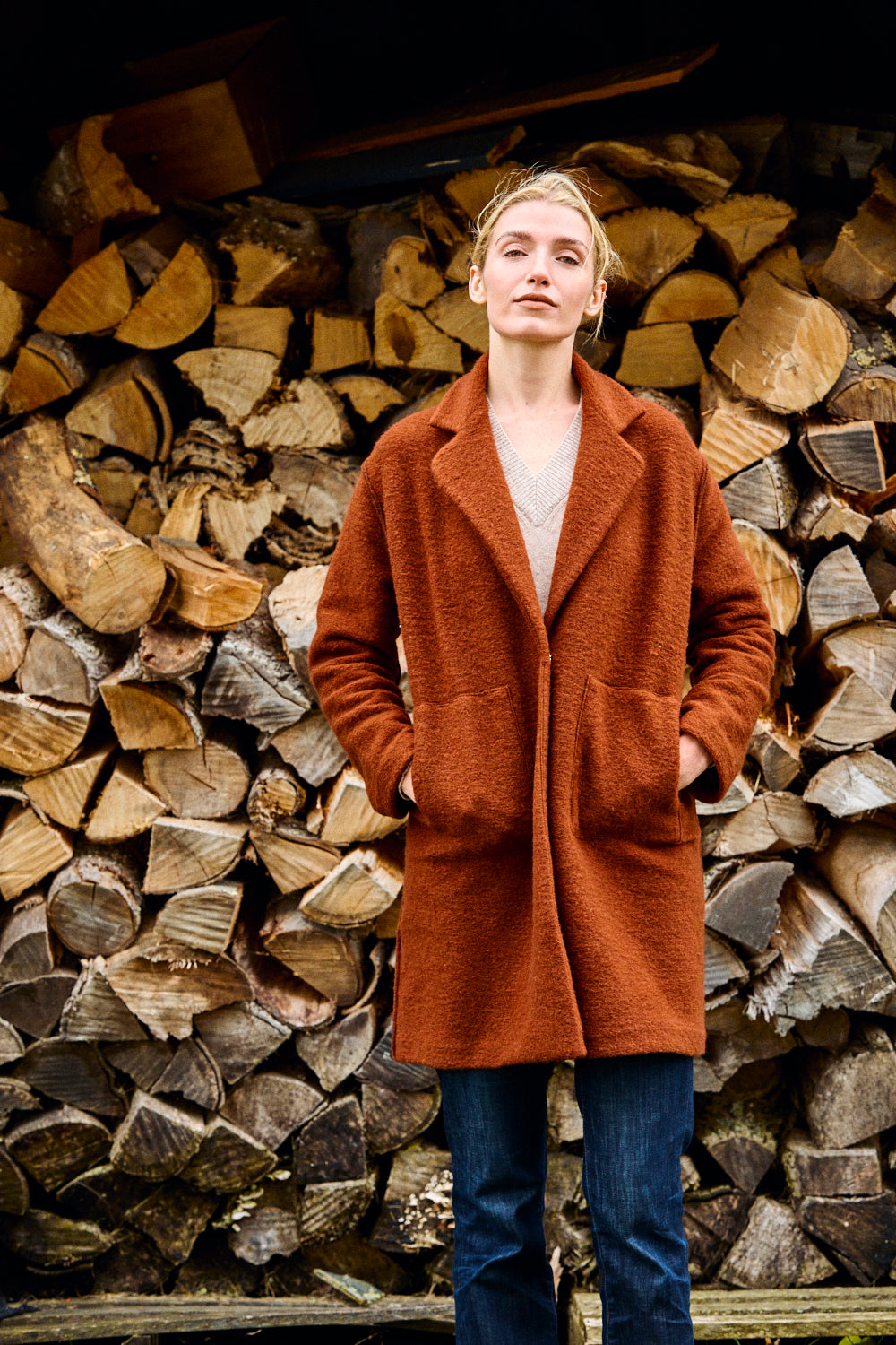 Person wearing a brown coat standing in front of stacked firewood