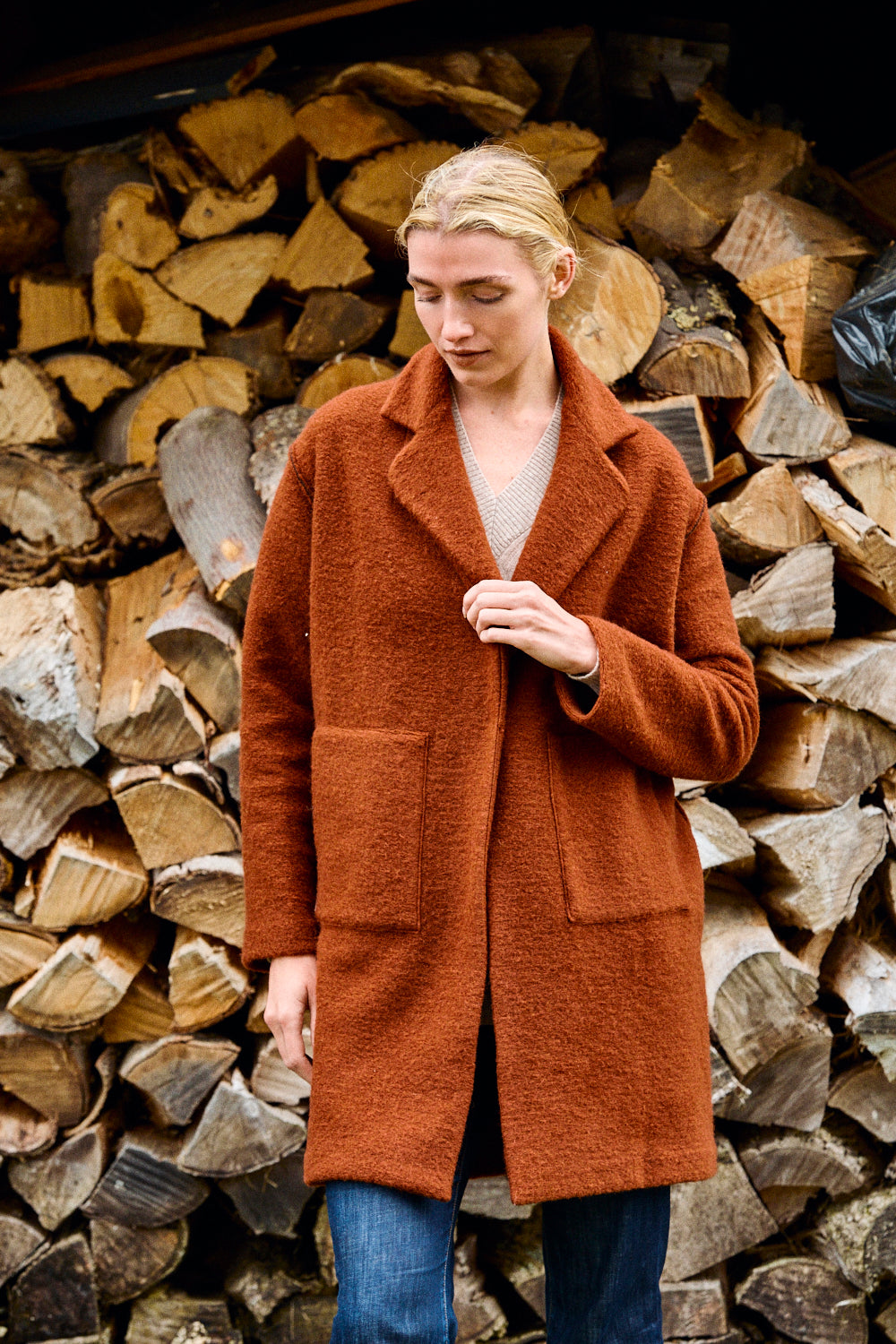 Woman wearing a brown coat standing in front of stacked firewood.