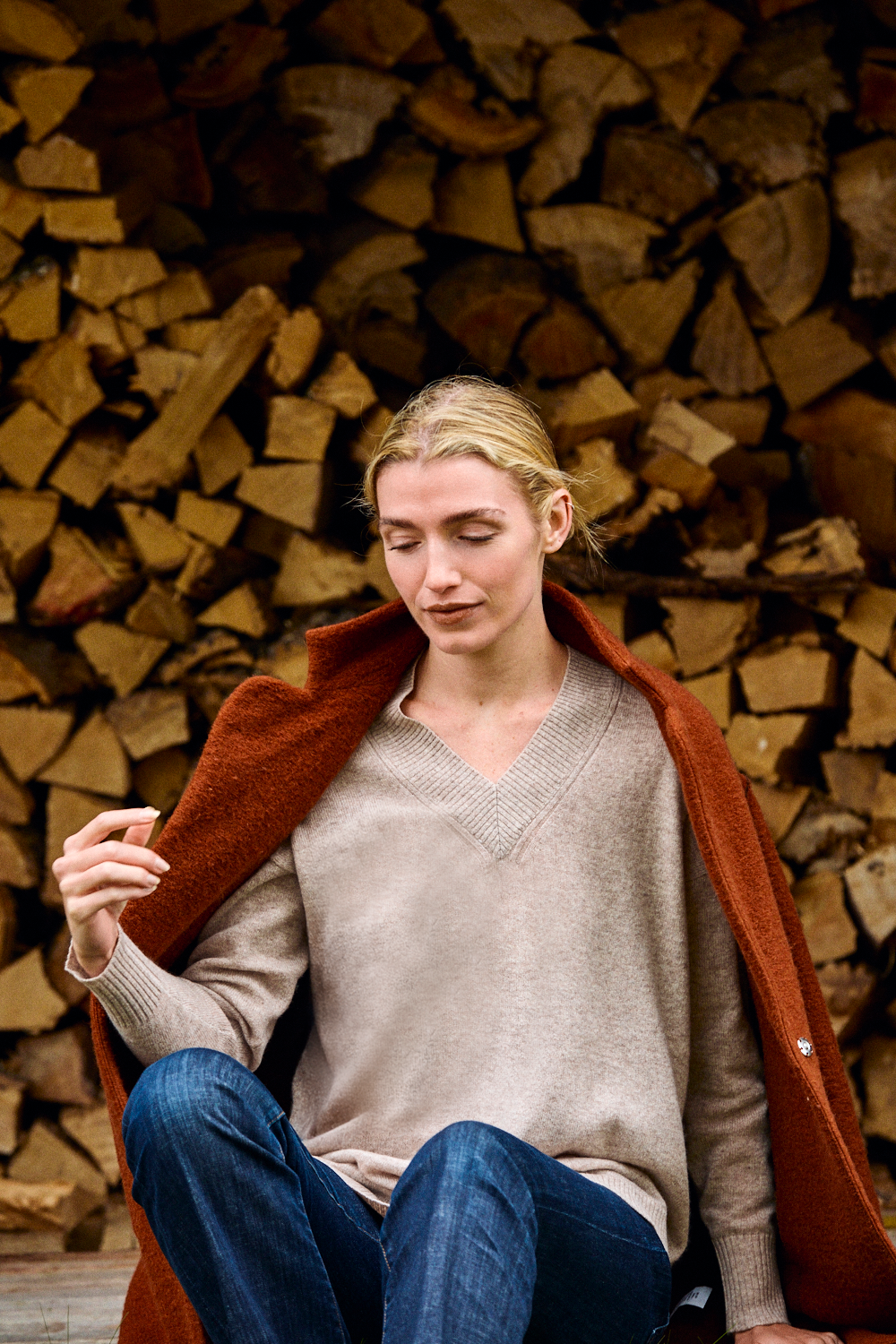 Woman sitting against a background of stacked firewood wearing a beige sweater and blue jeans.