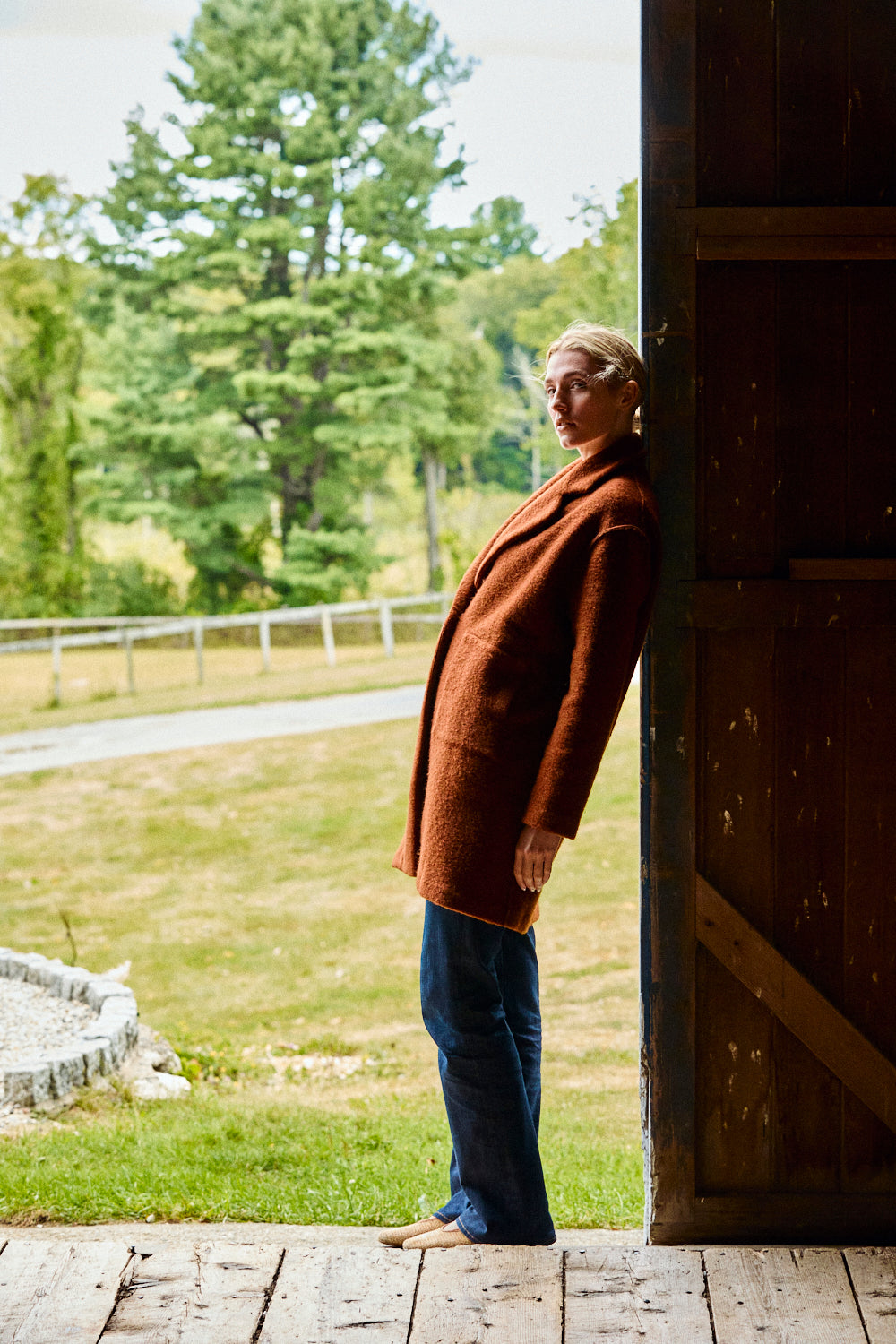 Person wearing a brown coat standing on a wooden deck with a grassy area and trees in the background.