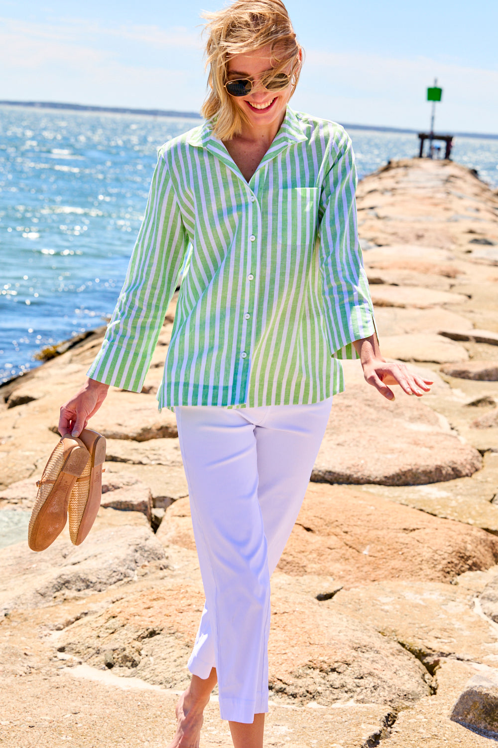 Woman in a striped shirt and white pants standing on a rocky beach.
