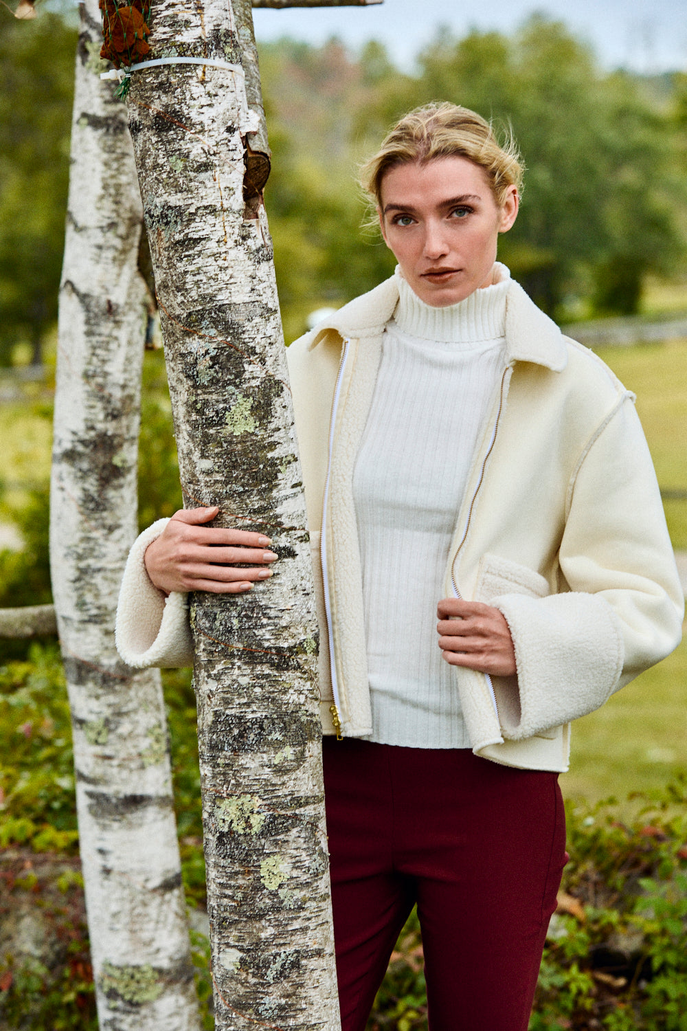 Woman in a cream coat and white sweater standing next to a tree in a natural setting