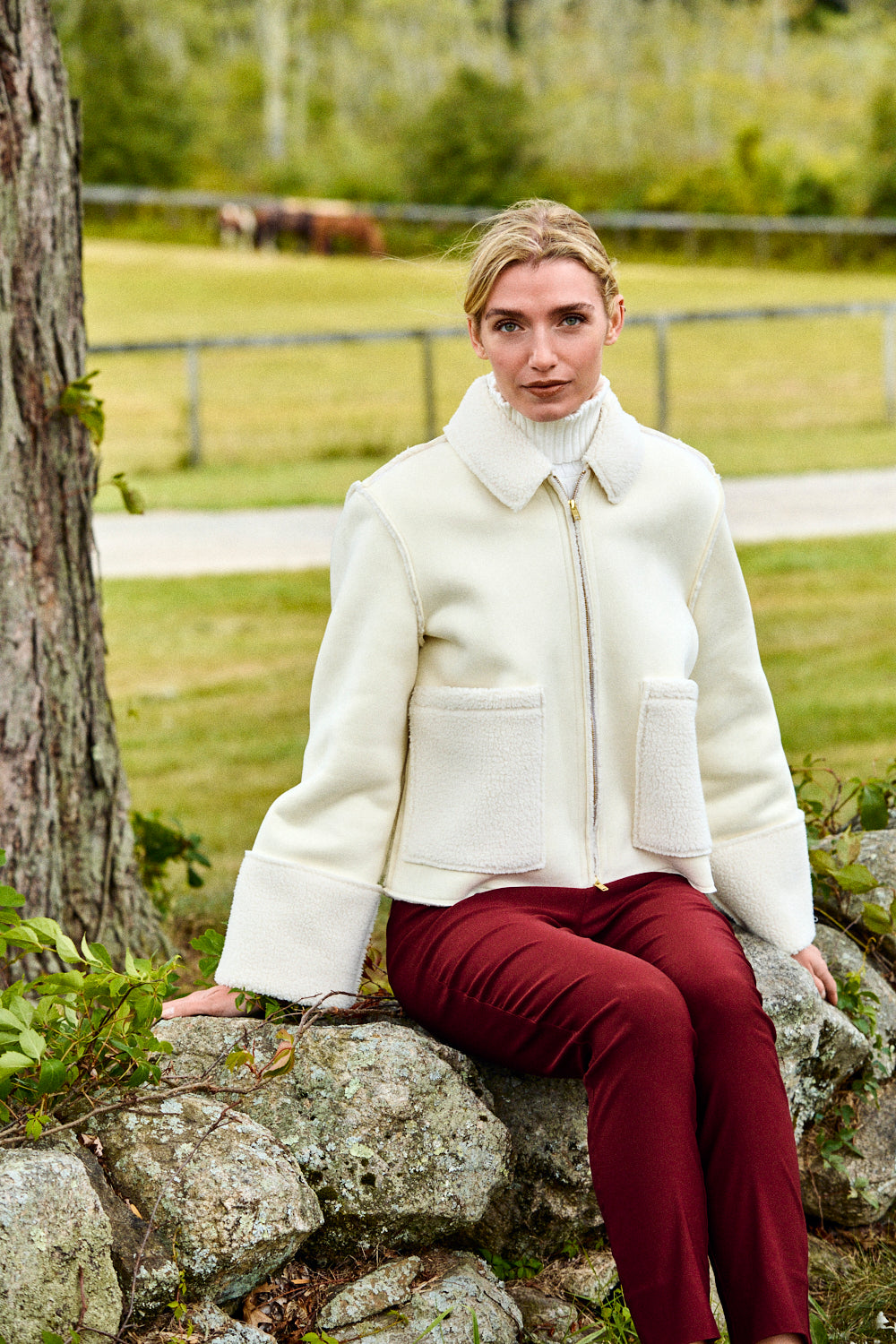 Woman in a white coat and red pants sitting on a rock with a horse in the background