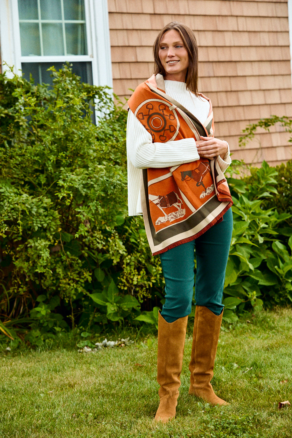 Woman wearing a patterned shawl, white sweater, blue jeans, and brown knee-high boots standing outdoors.