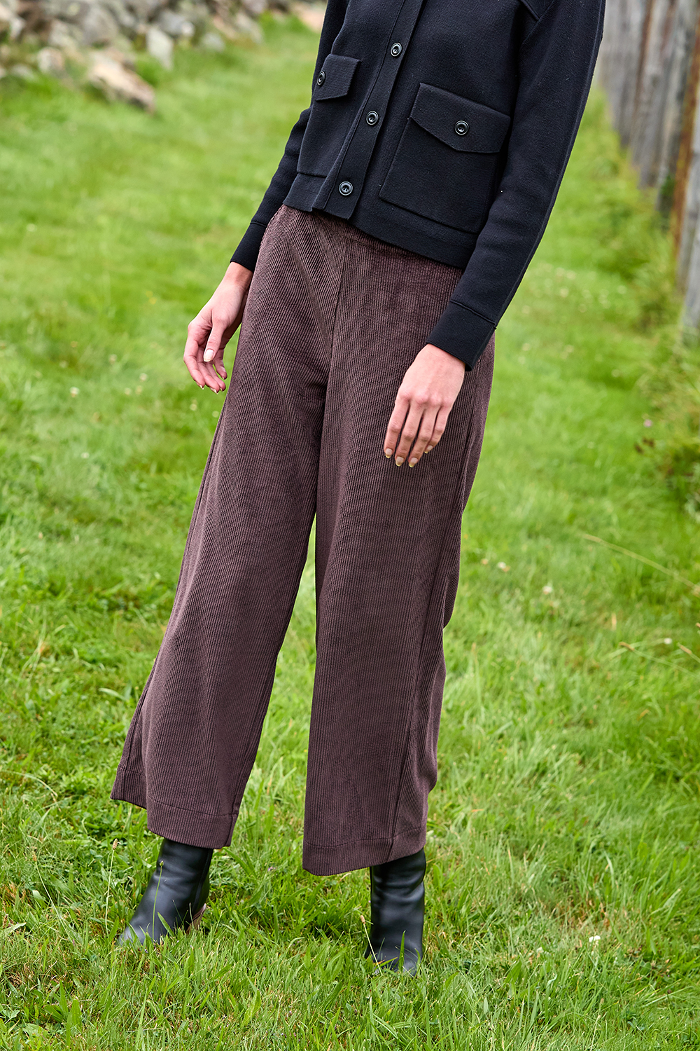 Person wearing brown wide-leg pants and black jacket standing in a grassy area.