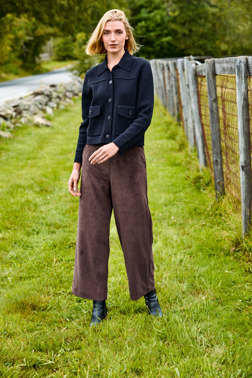 Woman standing in a grassy field wearing a dark jacket and brown pants.