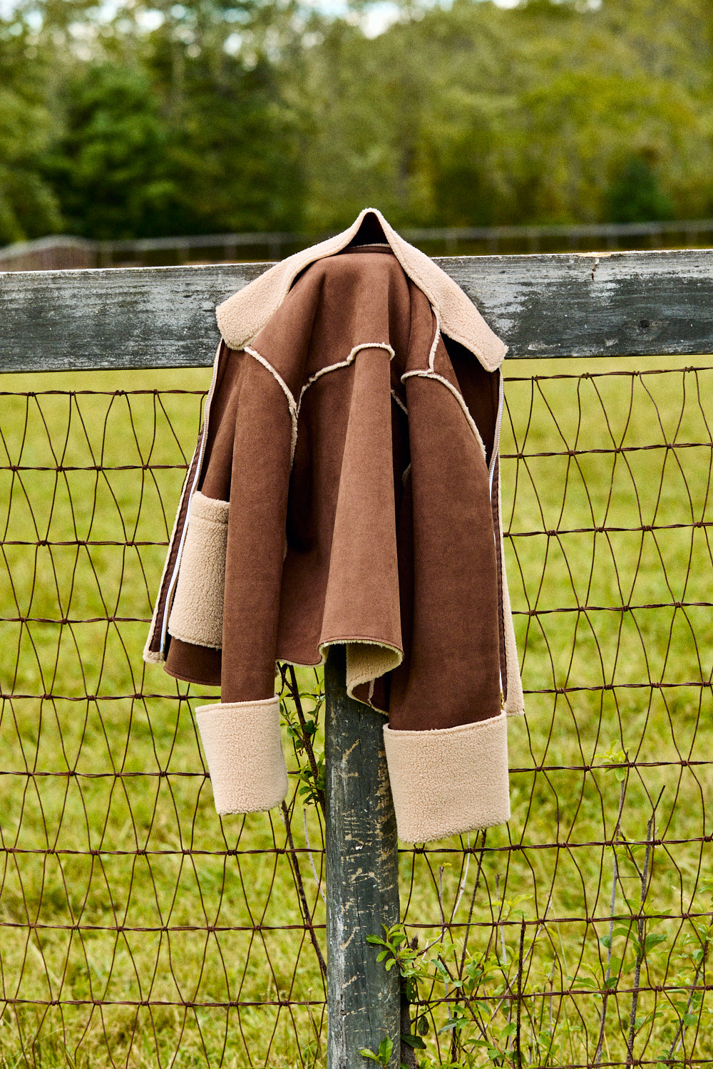 Brown and beige jacket hanging on a fence with a green field and trees in the background