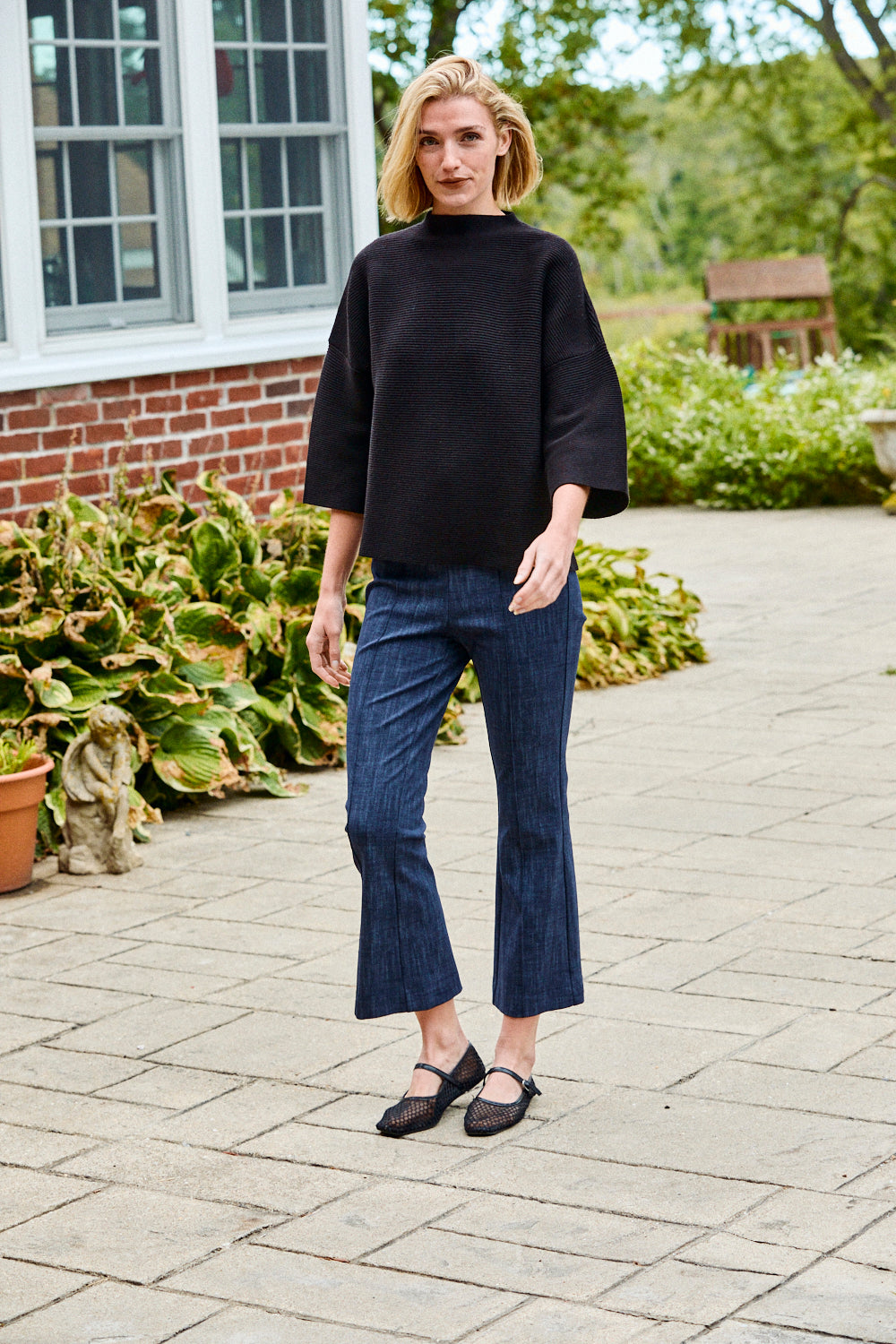 Woman wearing a black sweater and blue jeans standing on a paved walkway with greenery in the background.