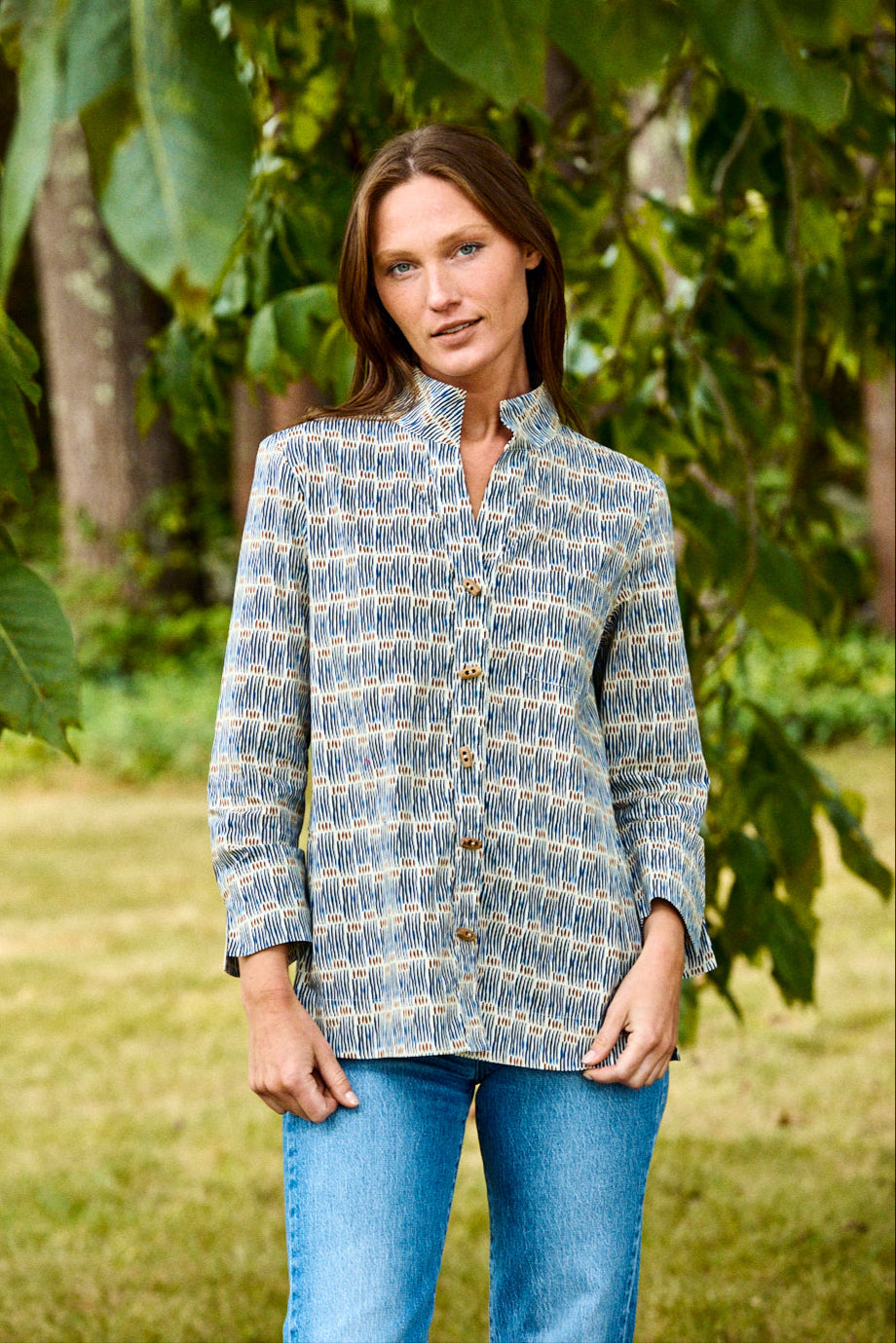 Woman wearing a blue patterned shirt and jeans standing in a natural setting with greenery.