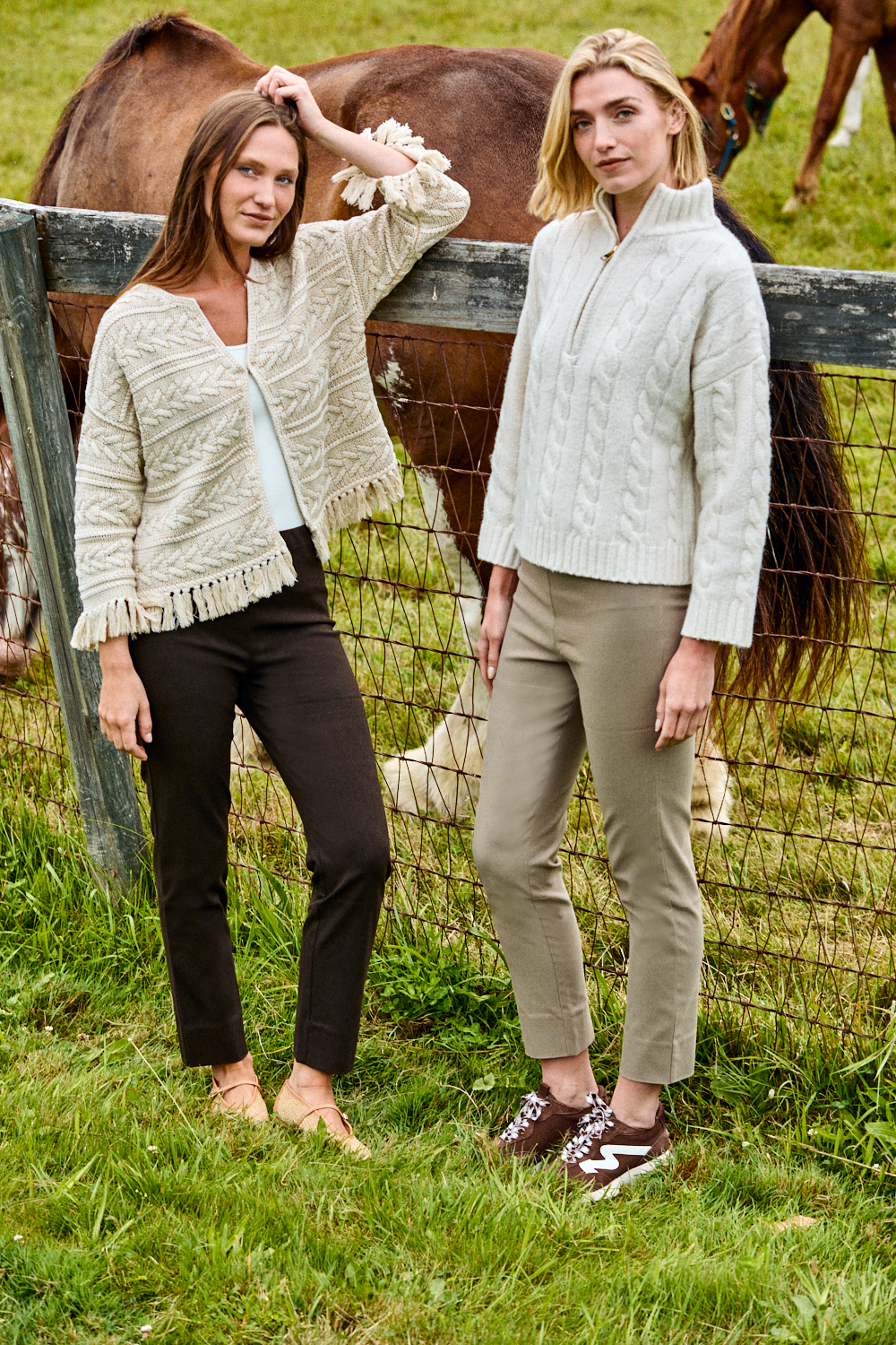 Two women wearing slim fit, tailored pants while standing in front of a fence and a horse.