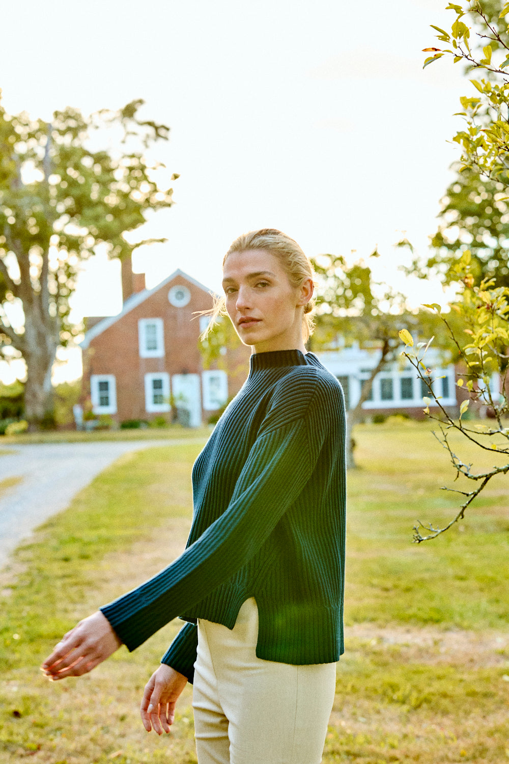 Woman in a green sweater standing outdoors with a house and trees in the background