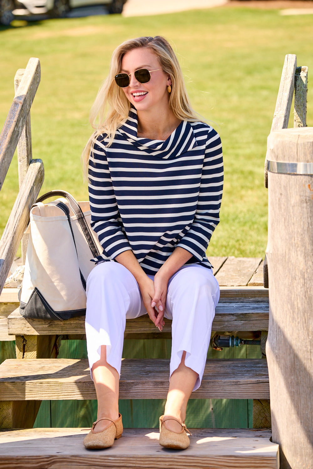 Woman sitting on a wooden bench wearing a striped shirt and white pants, with a grassy area in the background.