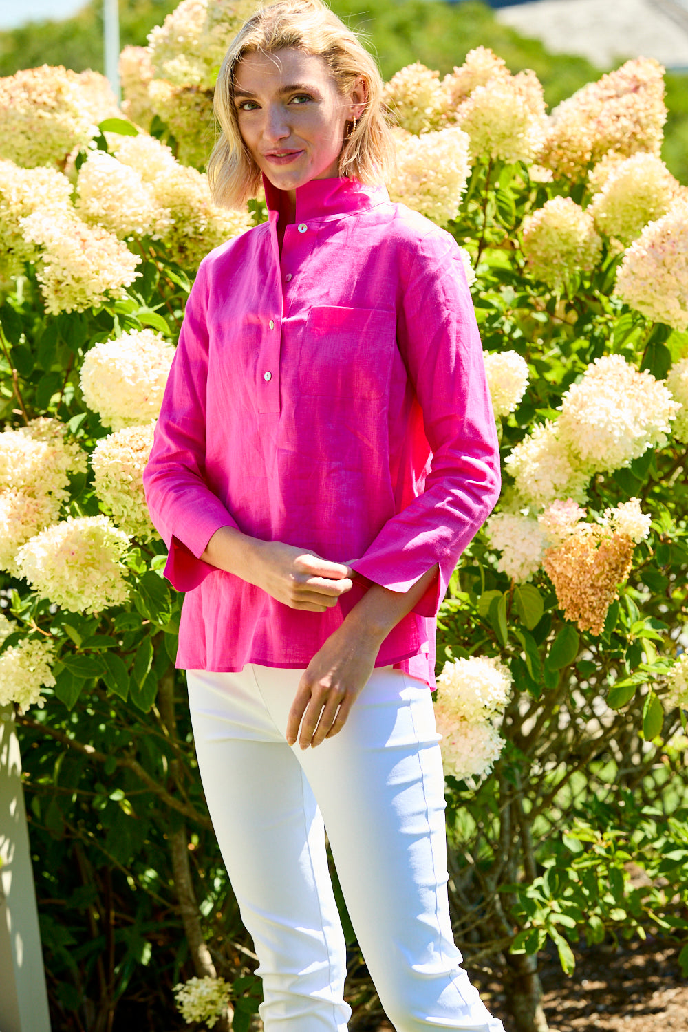 Woman wearing a bright pink shirt and white pants standing in front of a bush with white flowers.