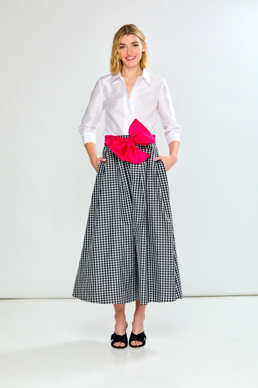 Woman wearing a white shirt, black and white checkered skirt with a pink bow, and black shoes on a white background.