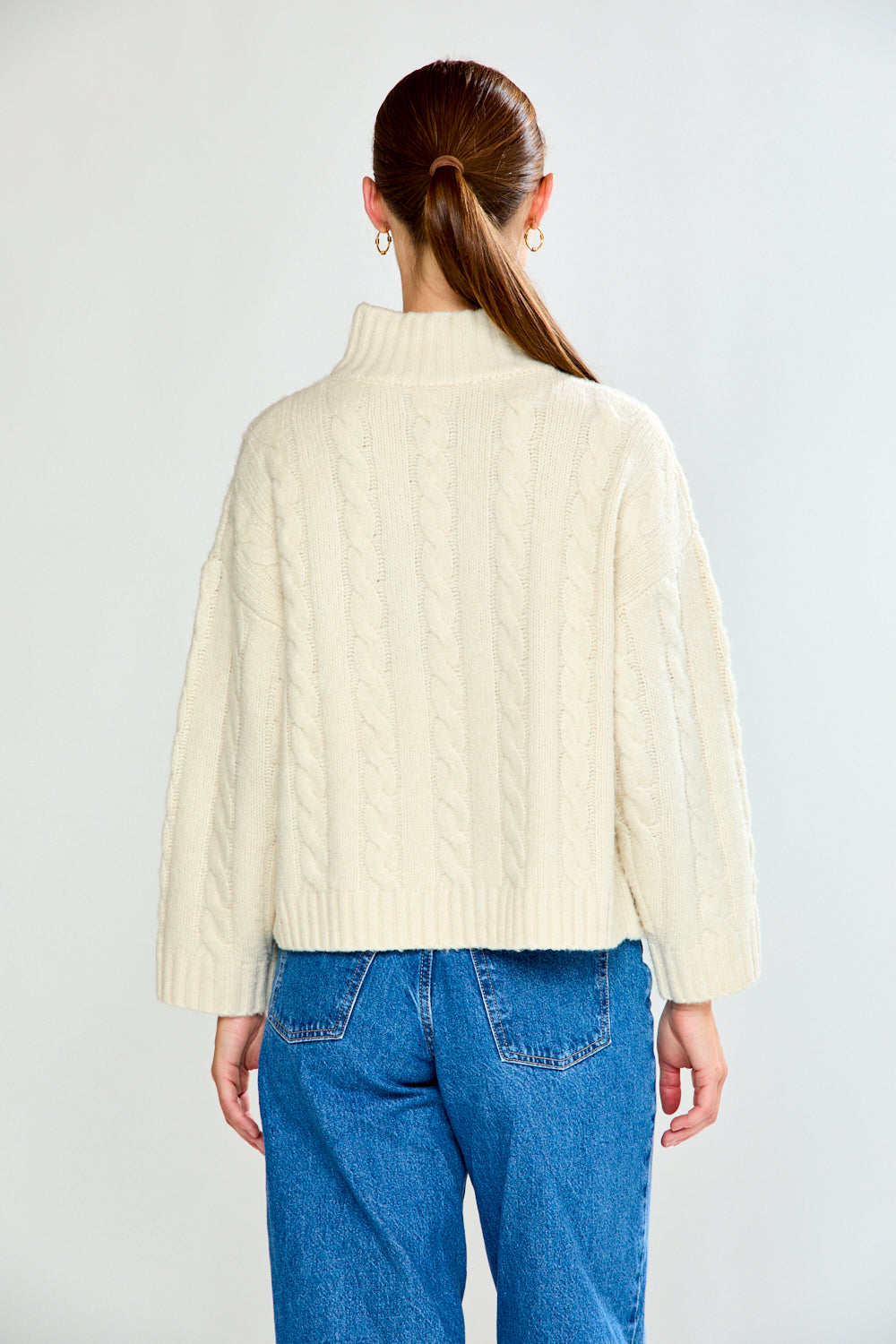 Woman wearing a cream cable knit sweater and blue jeans on a white background