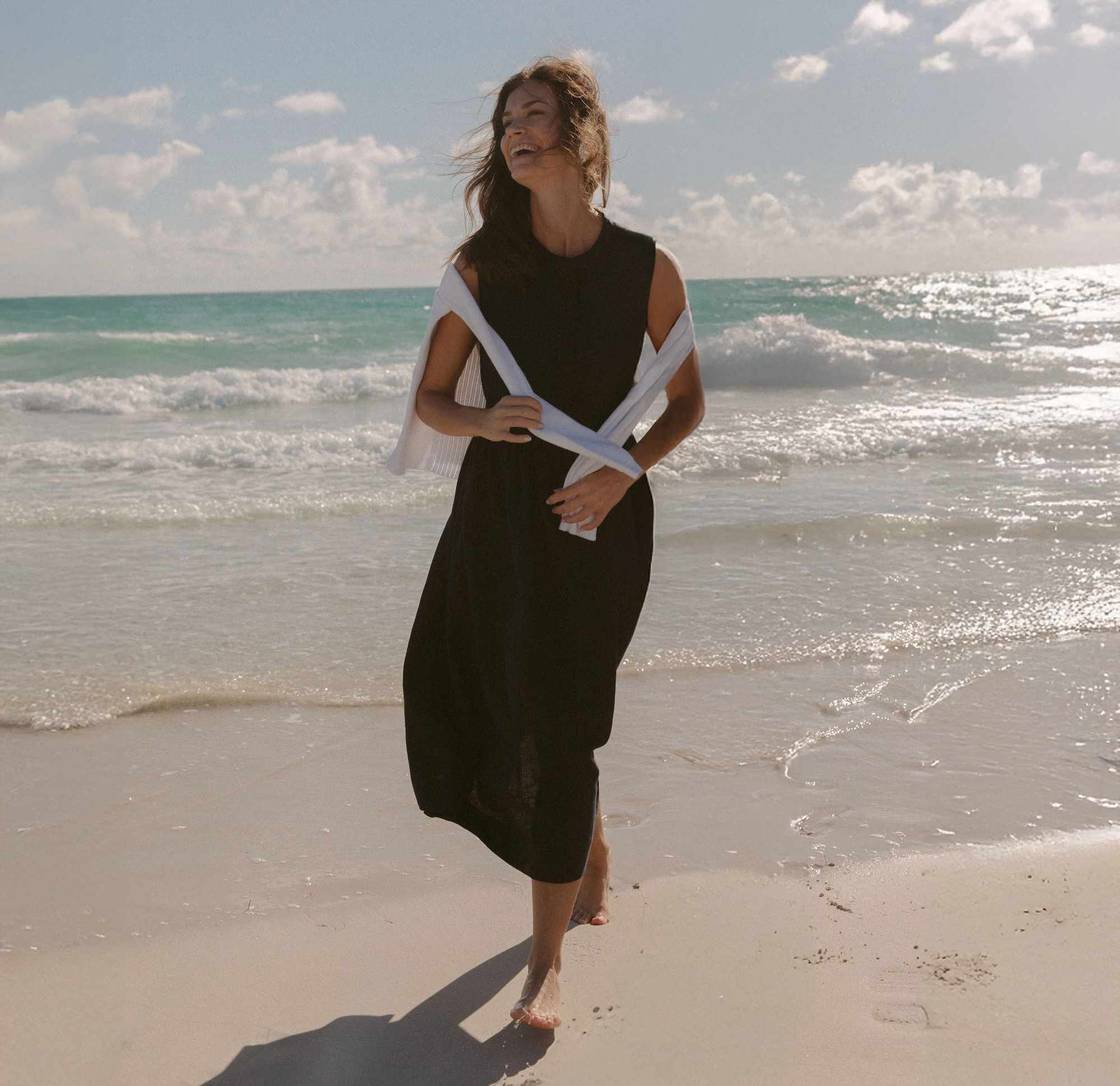 Woman wearing a navy linen dress and a white sweater draped over her shoulders while walking on the beach