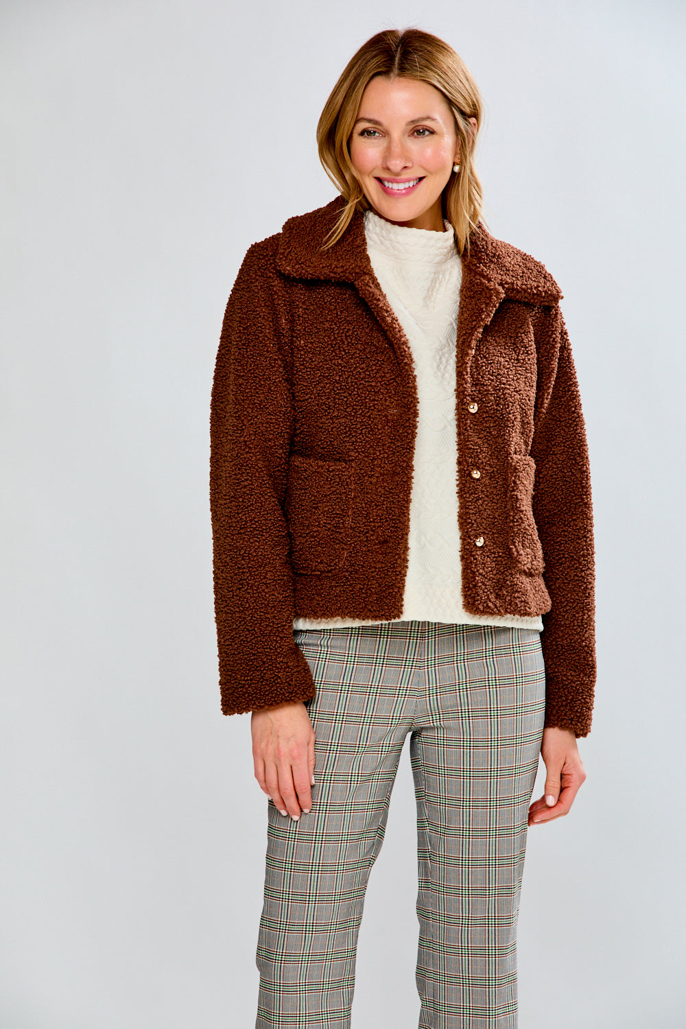 Woman wearing a brown teddy coat over a white sweater and plaid pants on a light gray background