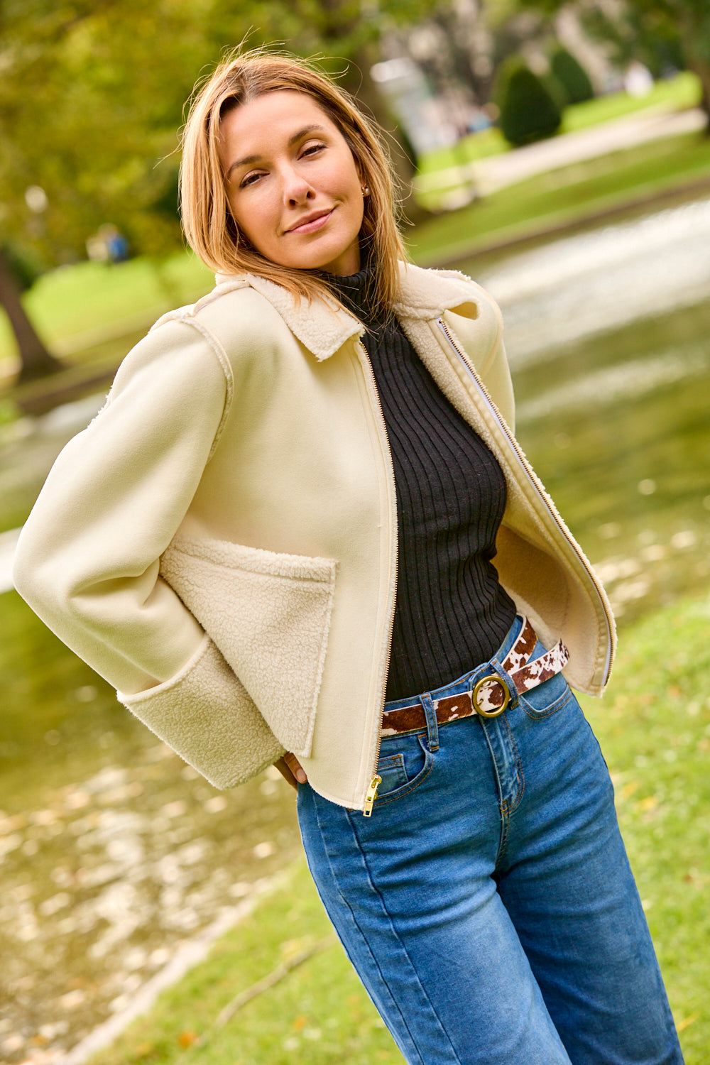 Woman wearing a white jacket, jeans, and a cowhide belt while standing outside in a park