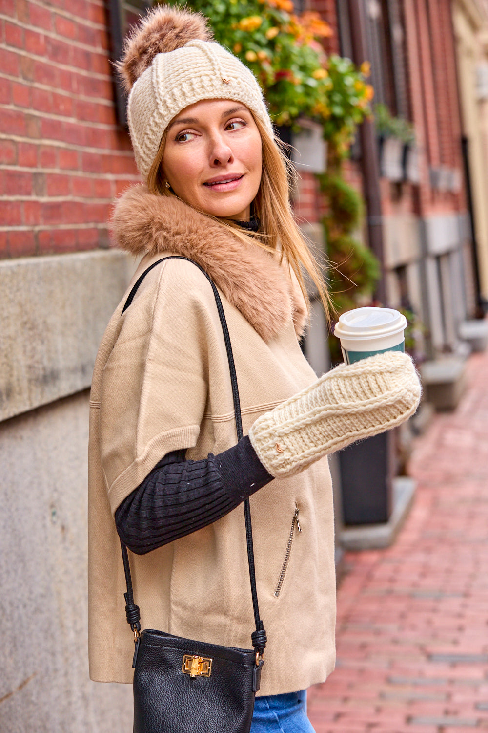 Woman wearing a short sleeve wheat colored sweater with a faux fur collar, while holding a coffee. 