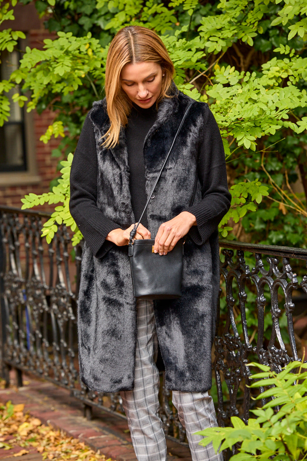Woman in a long black vest holding a black handbag outdoors with greenery in the background