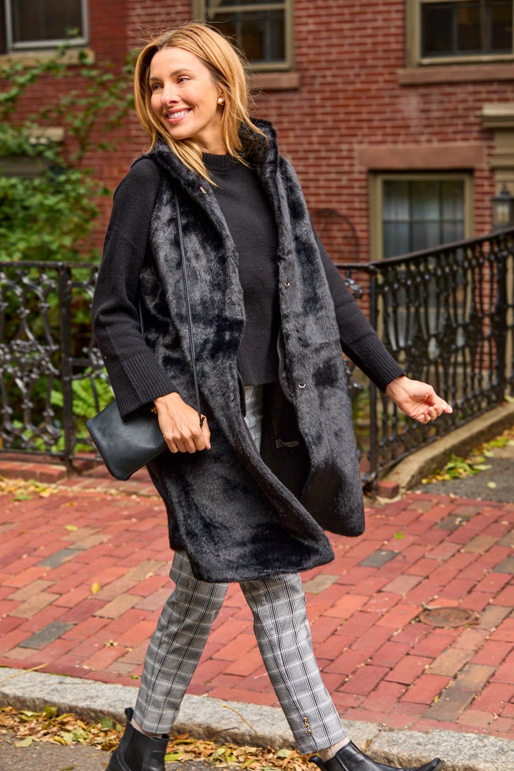 Woman walking outdoors wearing a black vest, black top, plaid pants, and black shoes.