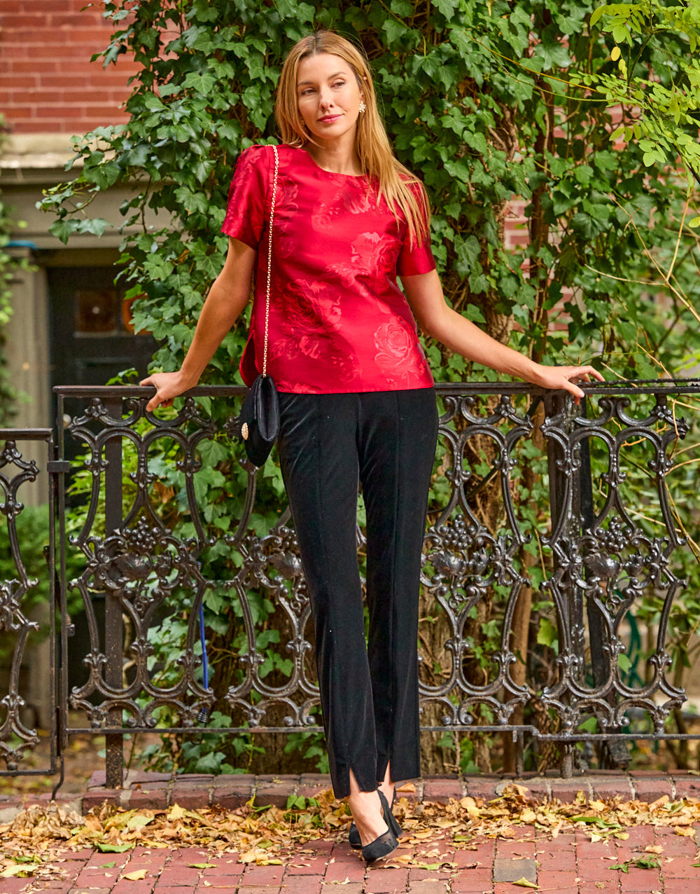 Woman wearing a red blouse and black pants while standing in front of a brick wall and iron fence. 