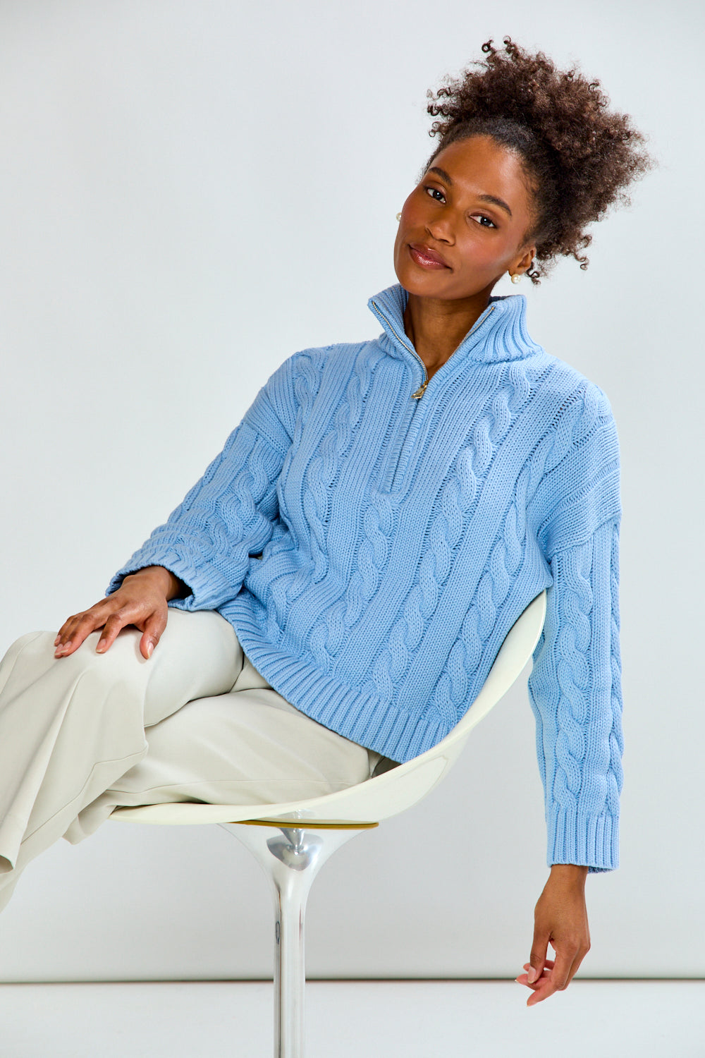 Woman wearing a light blue cable knit sweater sitting on a white chair against a white background