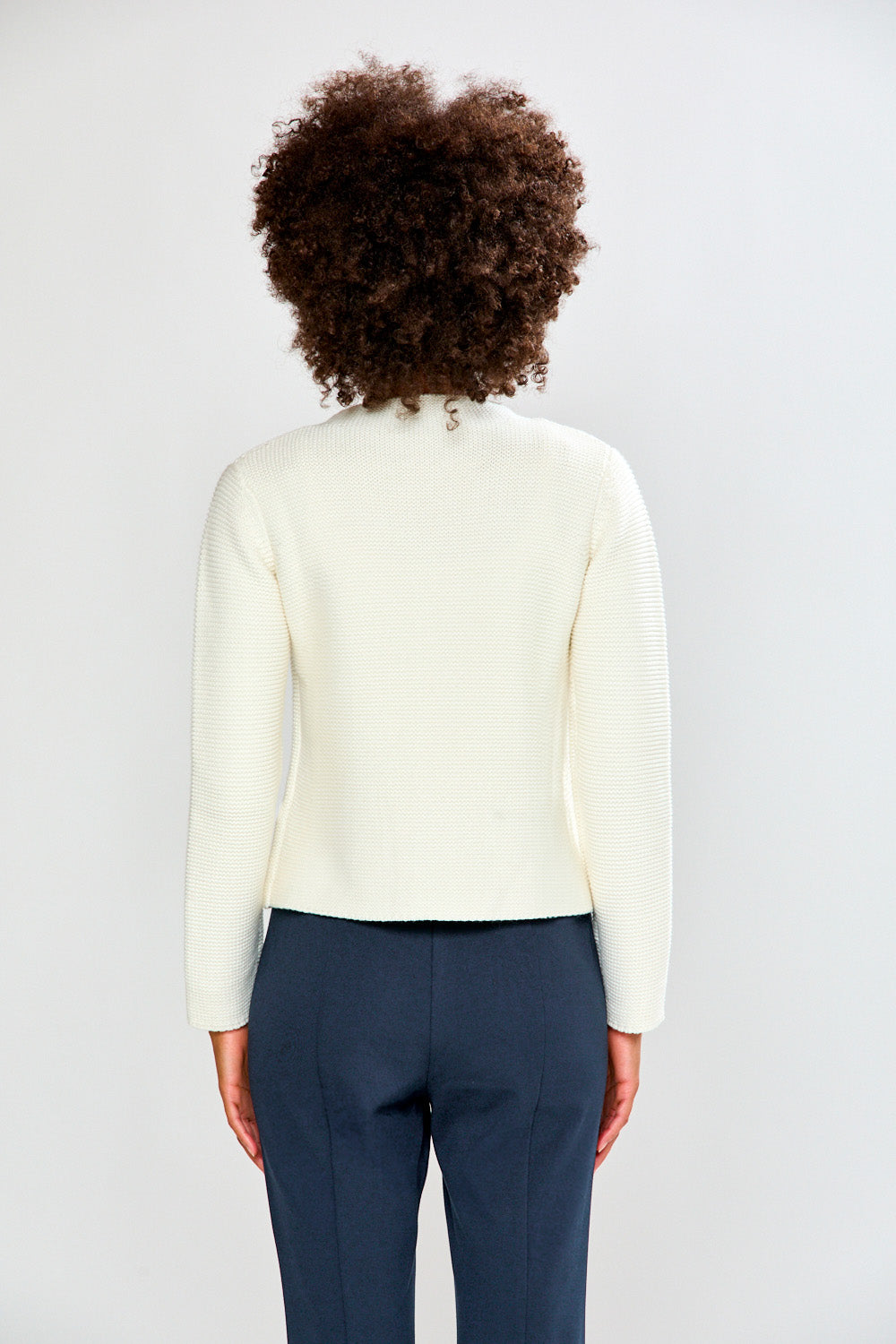 Woman wearing a white cardigan and navy pants against a plain background
