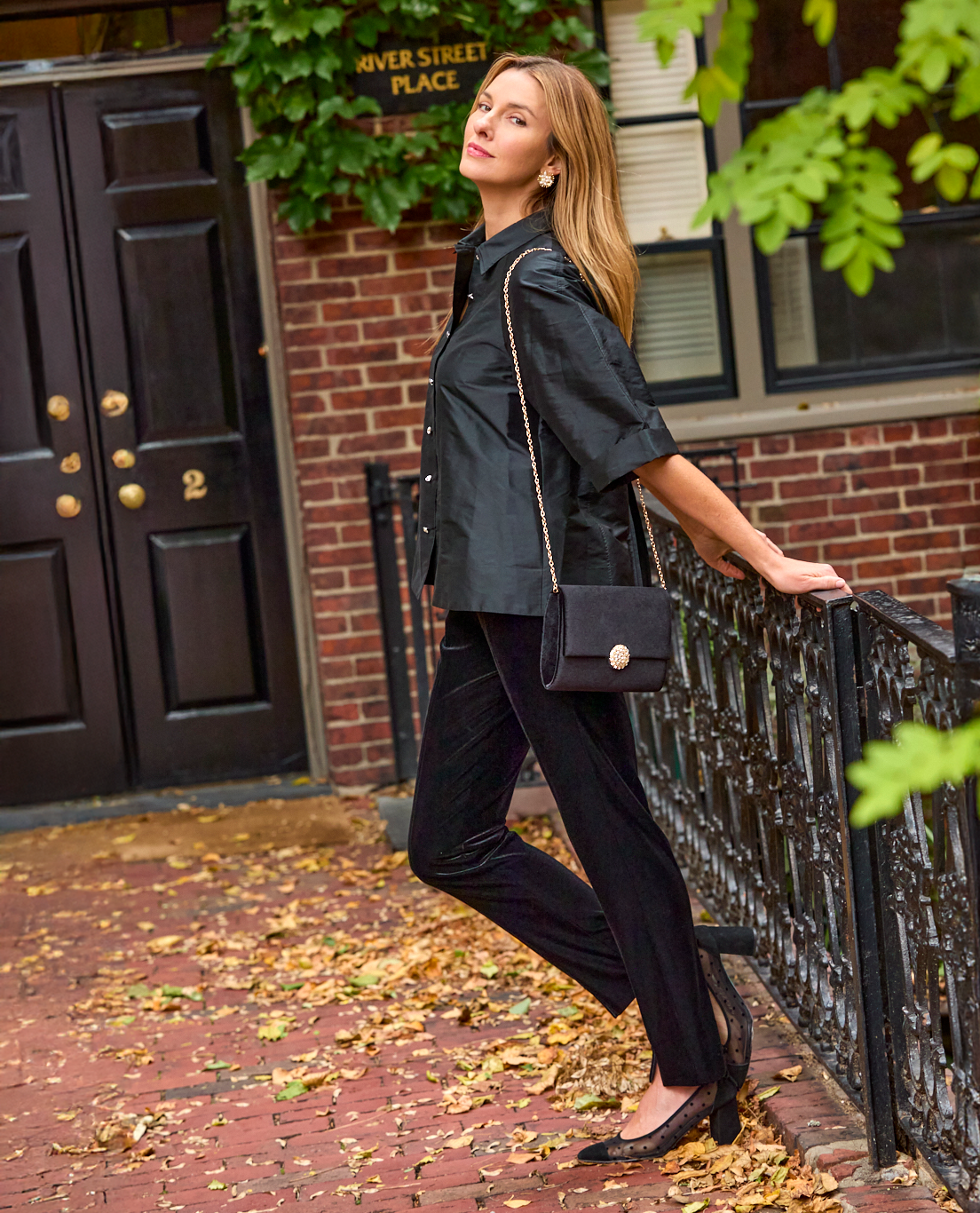 Woman wearing a black taffeta blouse and black velvet pants while leaning on an iron gate on a city sidewalk