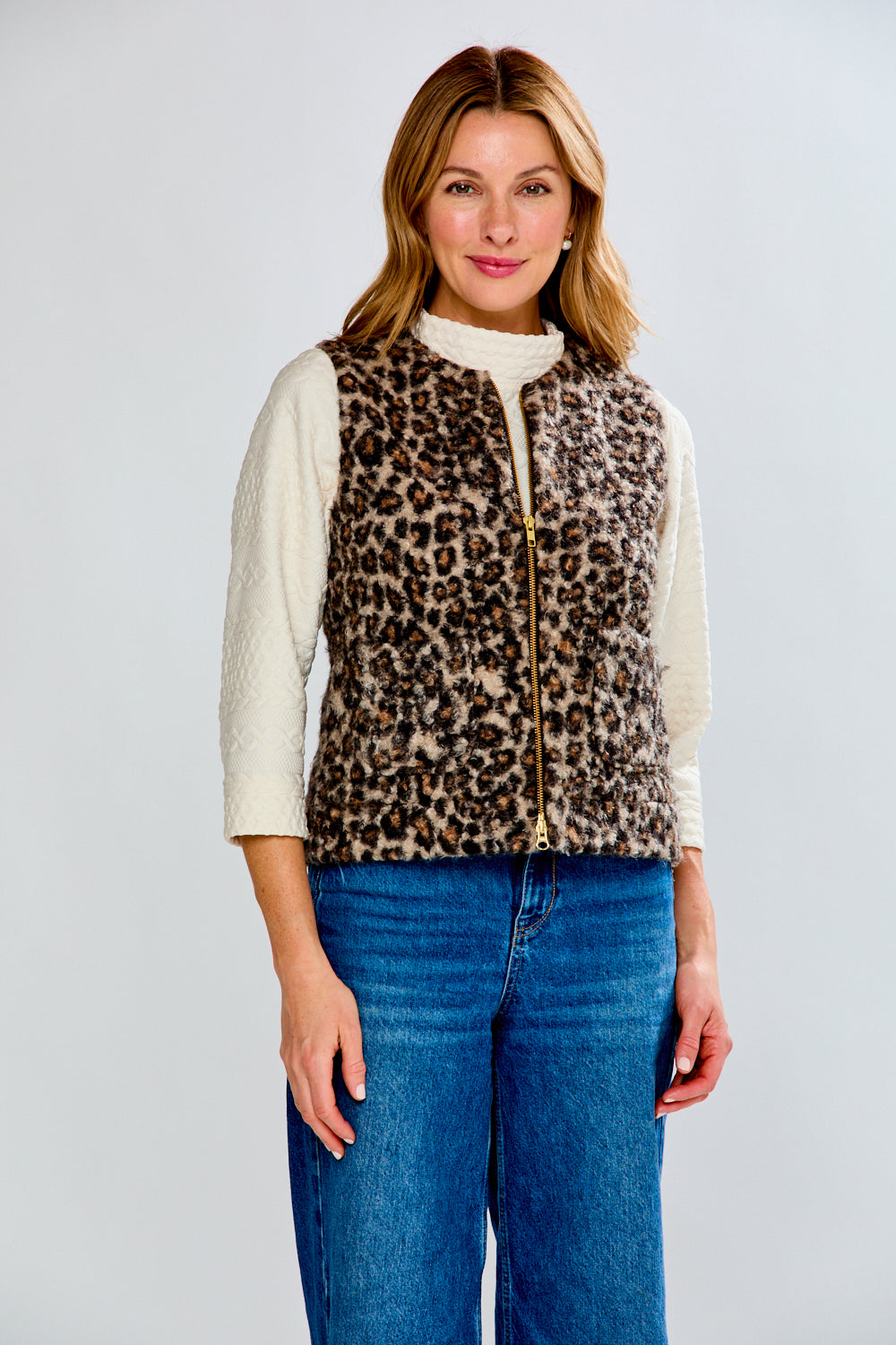 Woman wearing a leopard print vest over a white shirt with blue jeans on a light gray background