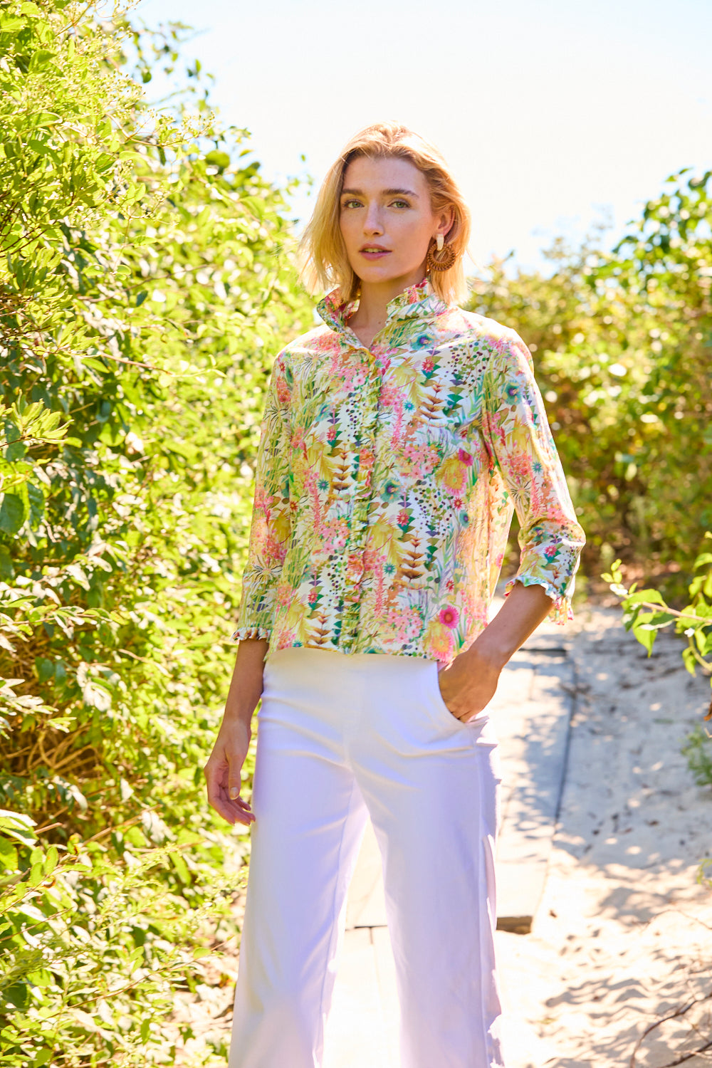 Woman wearing a floral blouse and white pants standing in a sunlit forest.