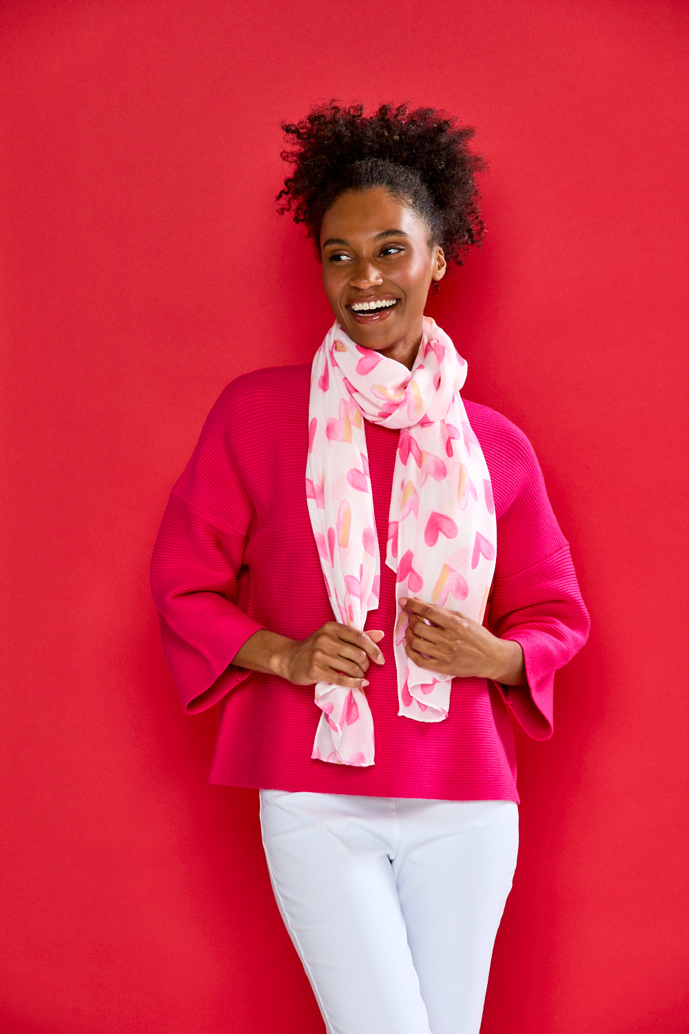 Woman wearing a pink sweater and white pants with a pink and white patterned scarf against a red background