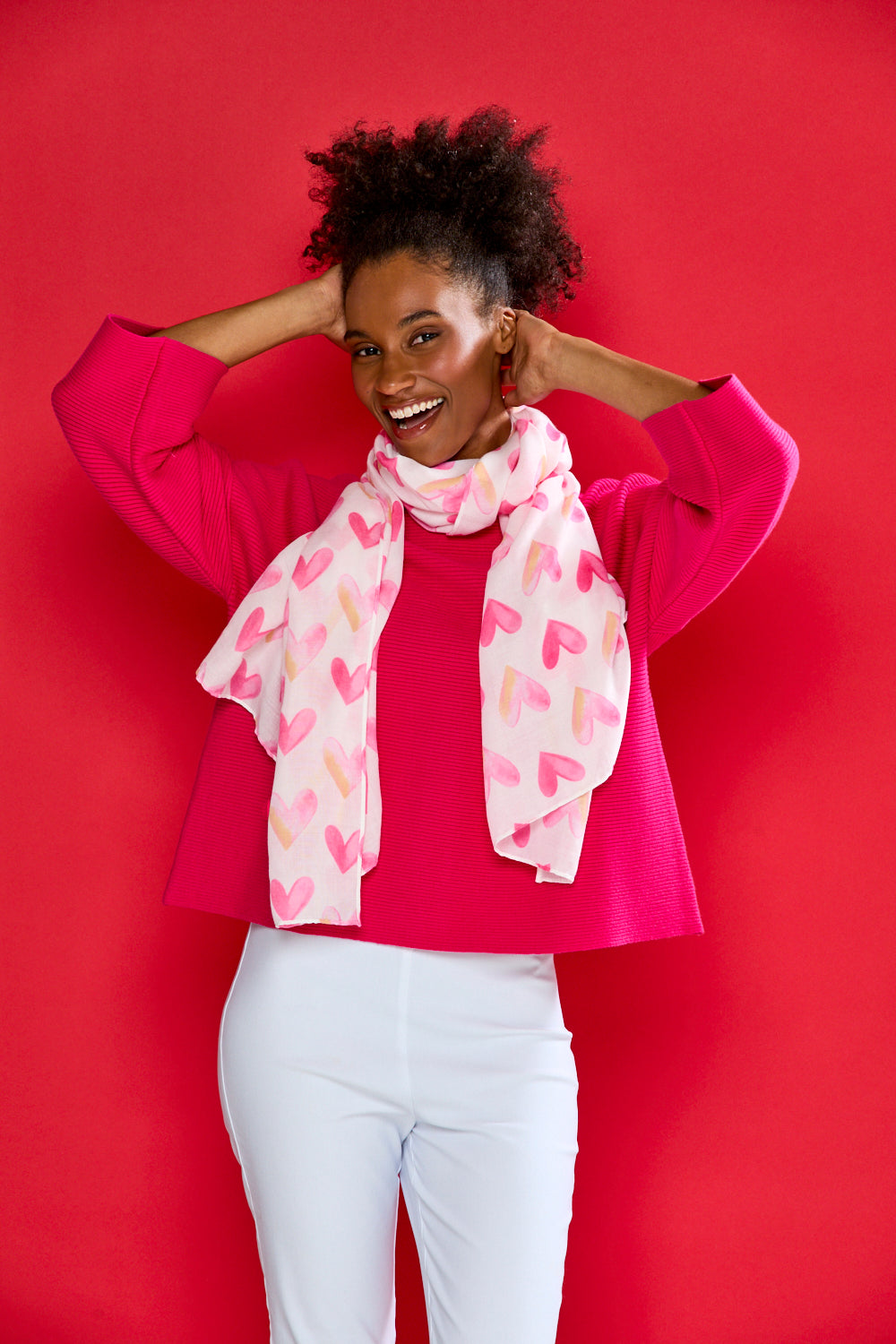 Woman wearing a pink jacket and white pants with a colorful scarf against a red background