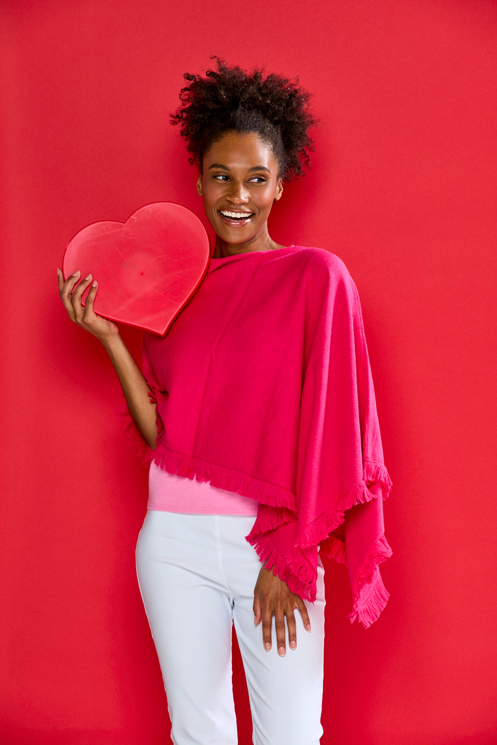 Woman wearing a pink cashmere ruana with fringe and holding a red heart in front of a red backdrop