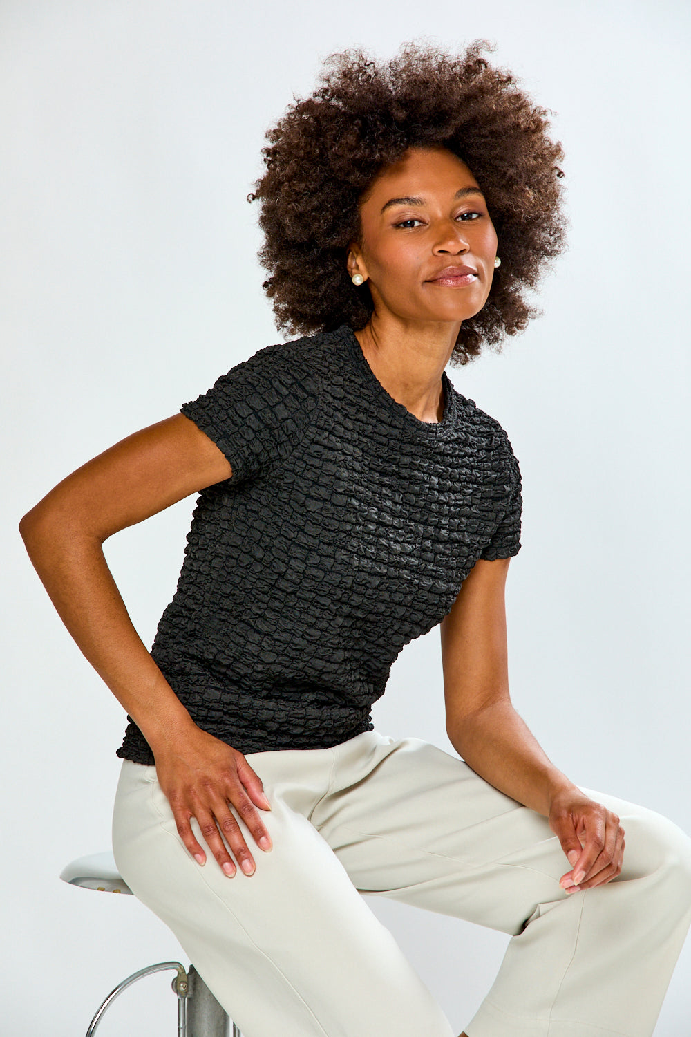 Woman wearing a textured black top and light-colored pants sitting on a white background