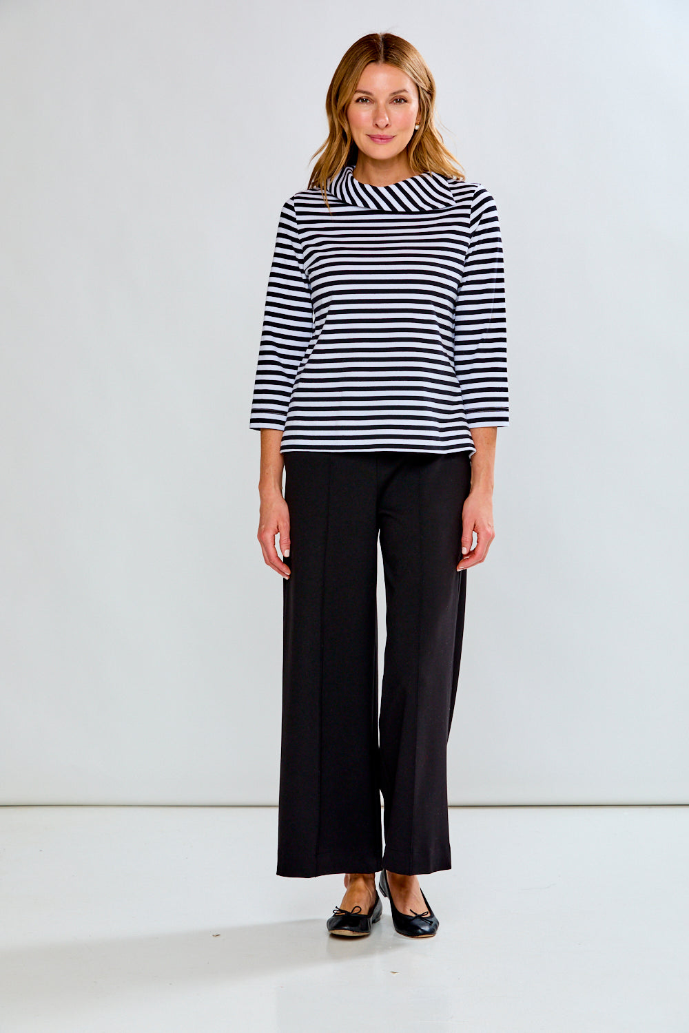 A woman modeling a three-quarter sleeve top with black and white horizontal stripes, paired with black wide-leg trousers and black flats, against a plain white studio background.