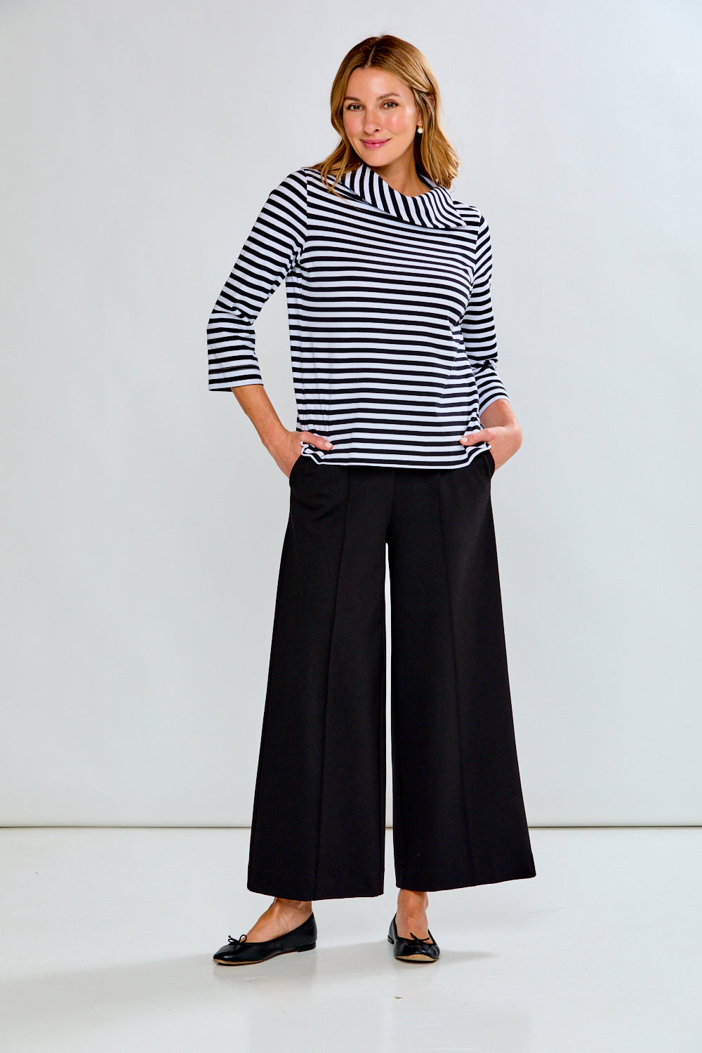 A woman modeling a three-quarter sleeve top with black and white horizontal stripes, paired with black wide-leg trousers and black flats, against a plain white studio background.