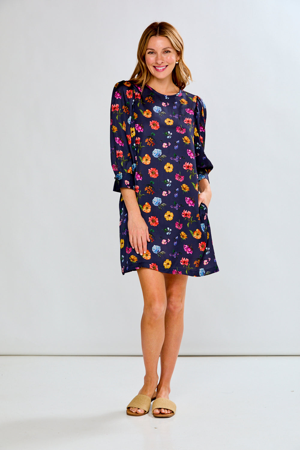 A woman modeling a navy blue short-sleeve shift dress with a colorful floral print, paired with tan slide sandals, against a plain white studio background.