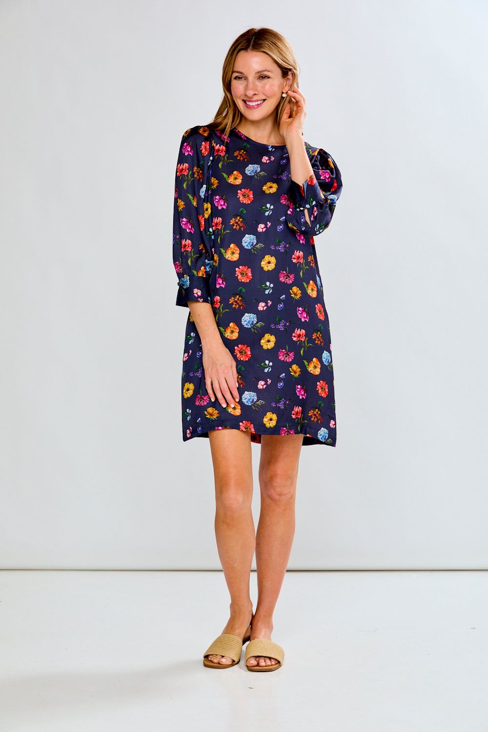 A woman modeling a navy blue short-sleeve shift dress with a colorful floral print, paired with tan slide sandals, against a plain white studio background.