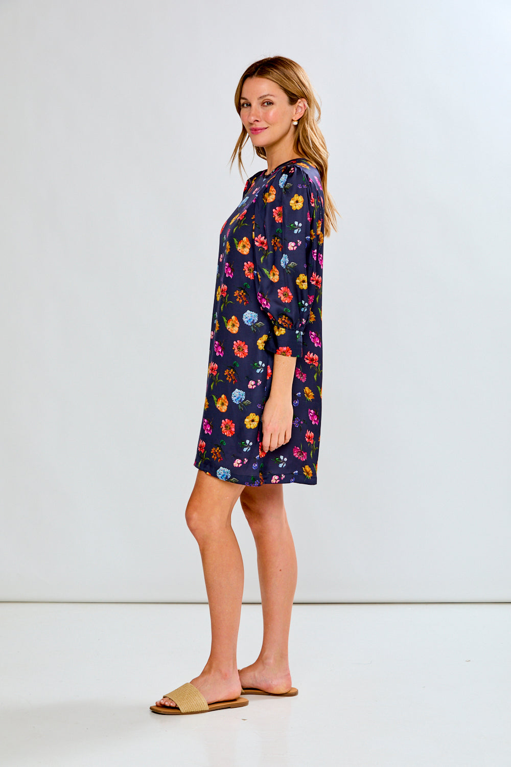 A woman modeling a navy blue short-sleeve shift dress with a colorful floral print, paired with tan slide sandals, against a plain white studio background.