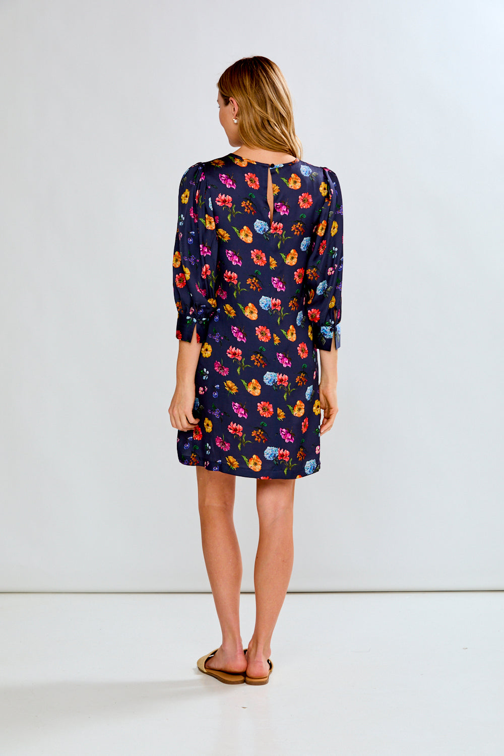 A woman modeling a navy blue short-sleeve shift dress with a colorful floral print, paired with tan slide sandals, against a plain white studio background.