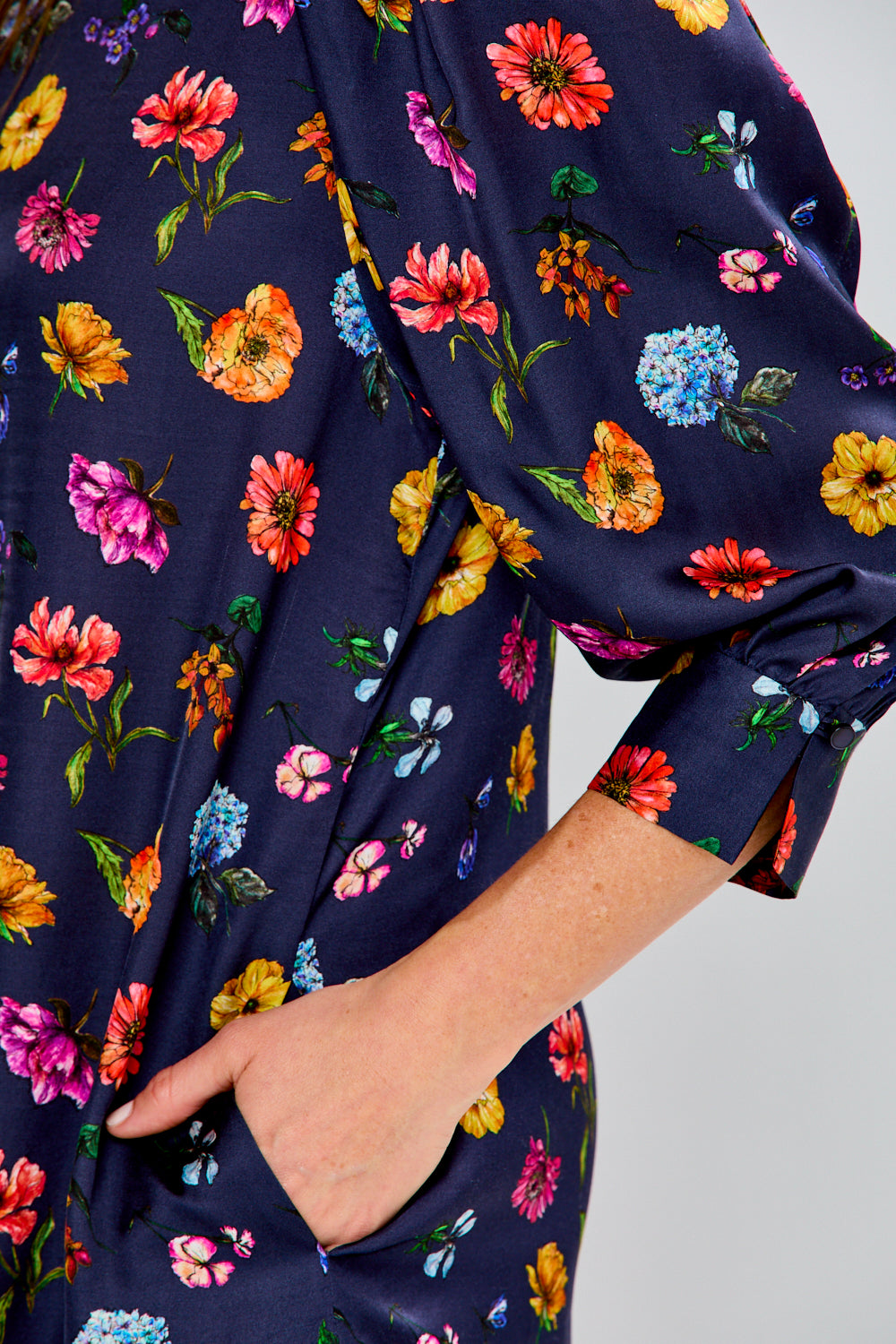 A woman modeling a navy blue short-sleeve shift dress with a colorful floral print, paired with tan slide sandals, against a plain white studio background.
