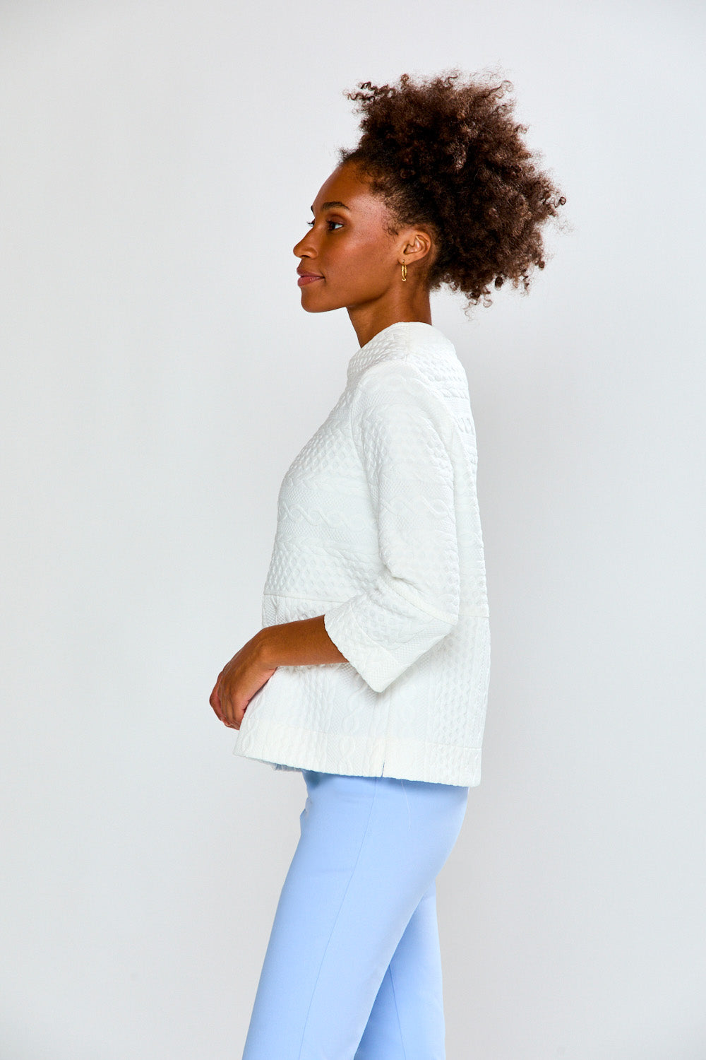 Woman wearing a white sweater and light blue pants against a plain background