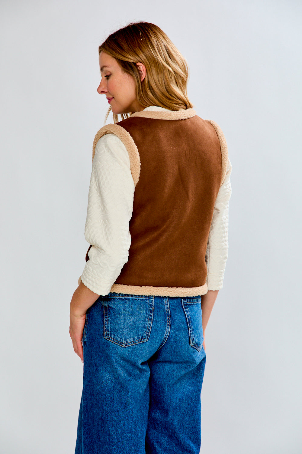 Person wearing a brown vest over a cream sweater with blue jeans on a light gray background