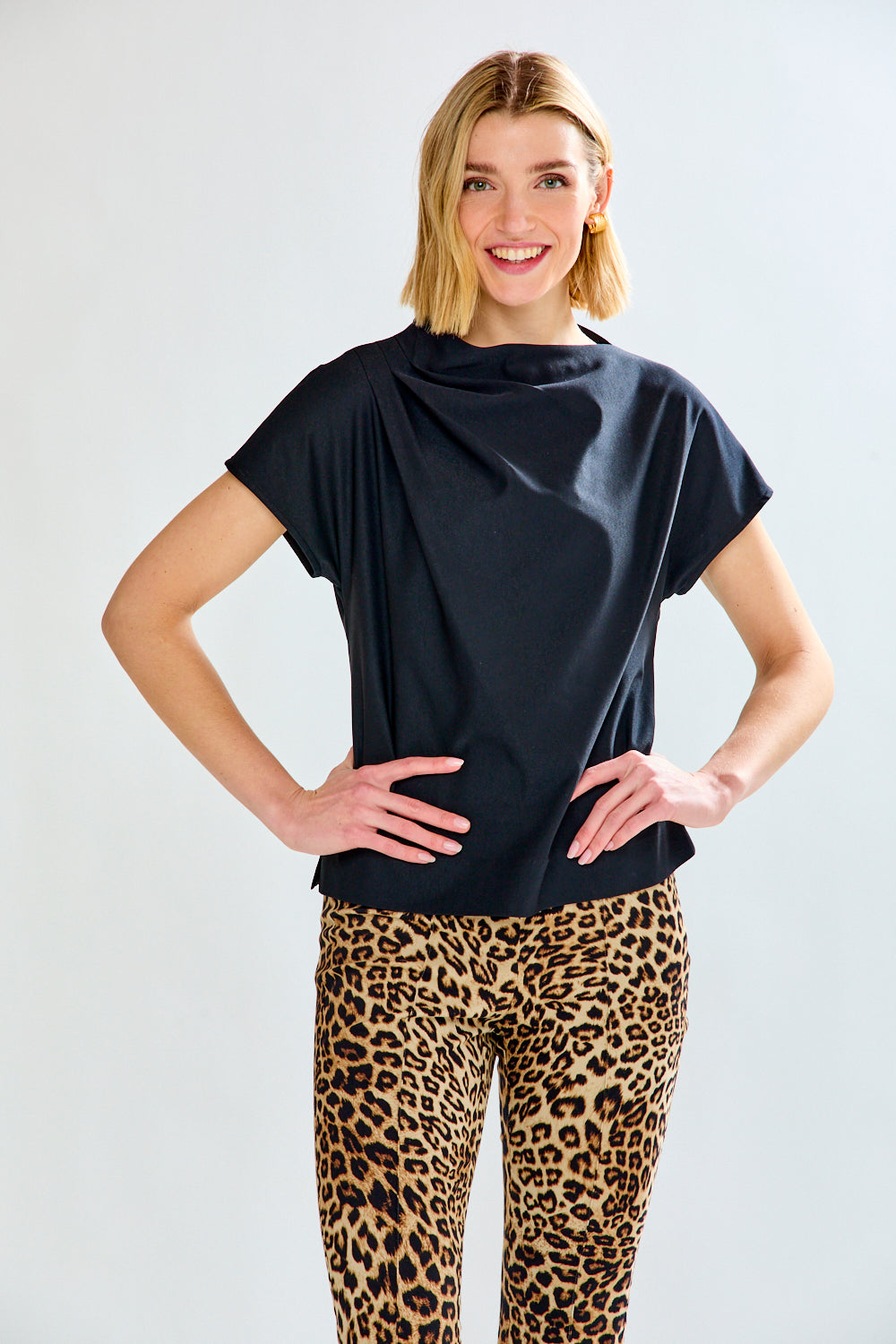 A woman modeling a black draped short-sleeve top with leopard-print pants against a light gray studio background.