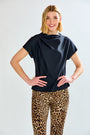 A woman modeling a black draped short-sleeve top with leopard-print pants against a light gray studio background.