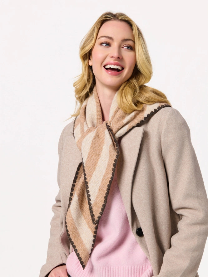 A woman wearing a beige and cream striped knit scarf with dark brown pom-pom trim, styled with a light beige cardigan and a pink sweater against a plain white background.